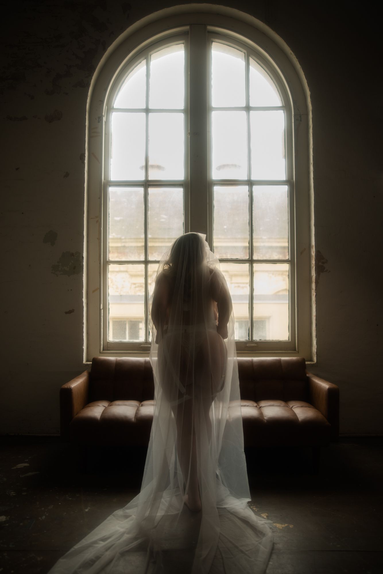 Woman in sheer fabric stands silhouetted before a large arched window. A brown sofa sits below the window in a dimly lit, aged room.