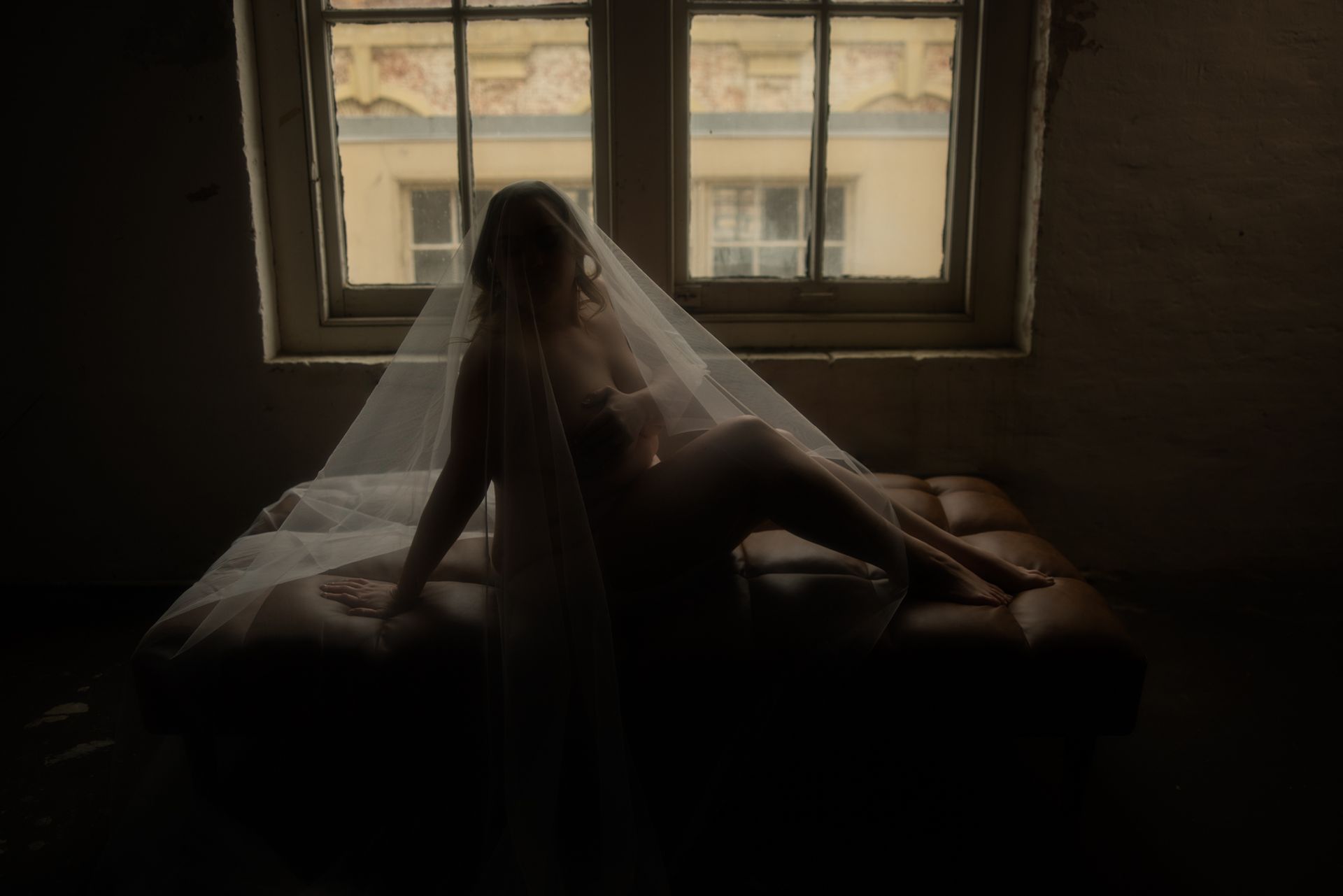 A nude woman draped in a sheer veil sits on a dark leather couch in front of a window, dimly lit.