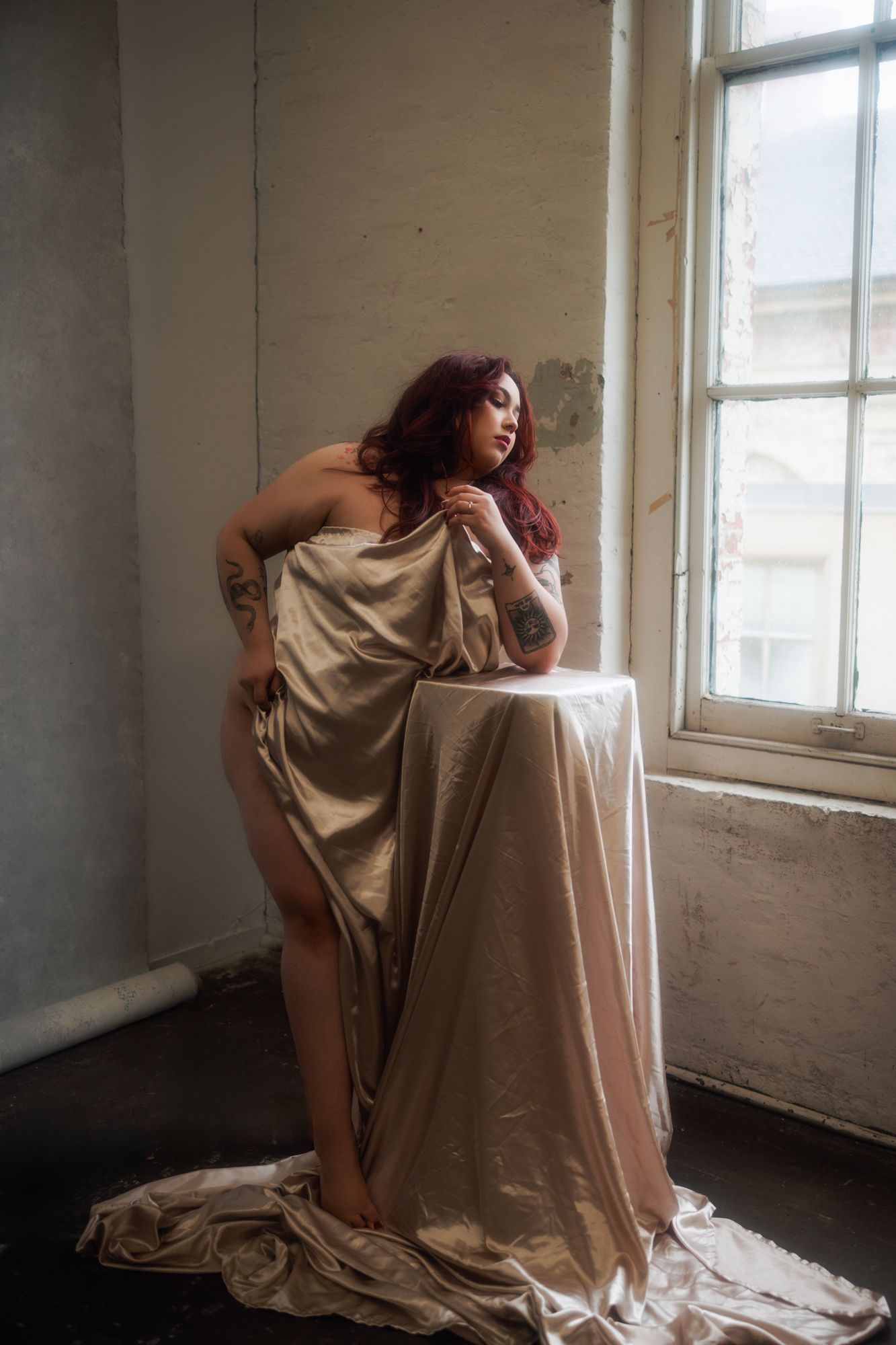 Woman with red hair draped in gold fabric, leaning on a table in front of a window in a room with peeling walls.