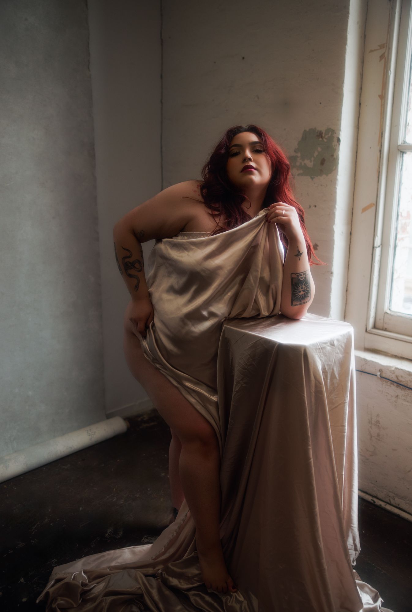 A woman with red hair draped in shimmering fabric poses beside a table in a room with a window.