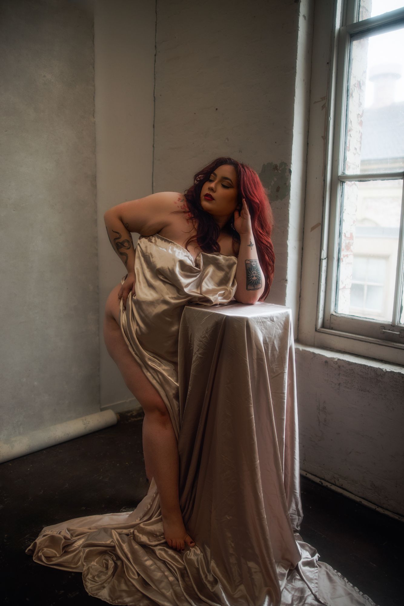 Woman with red hair draped in gold fabric, posing by a window. She leans on a small table, arms and legs visible.