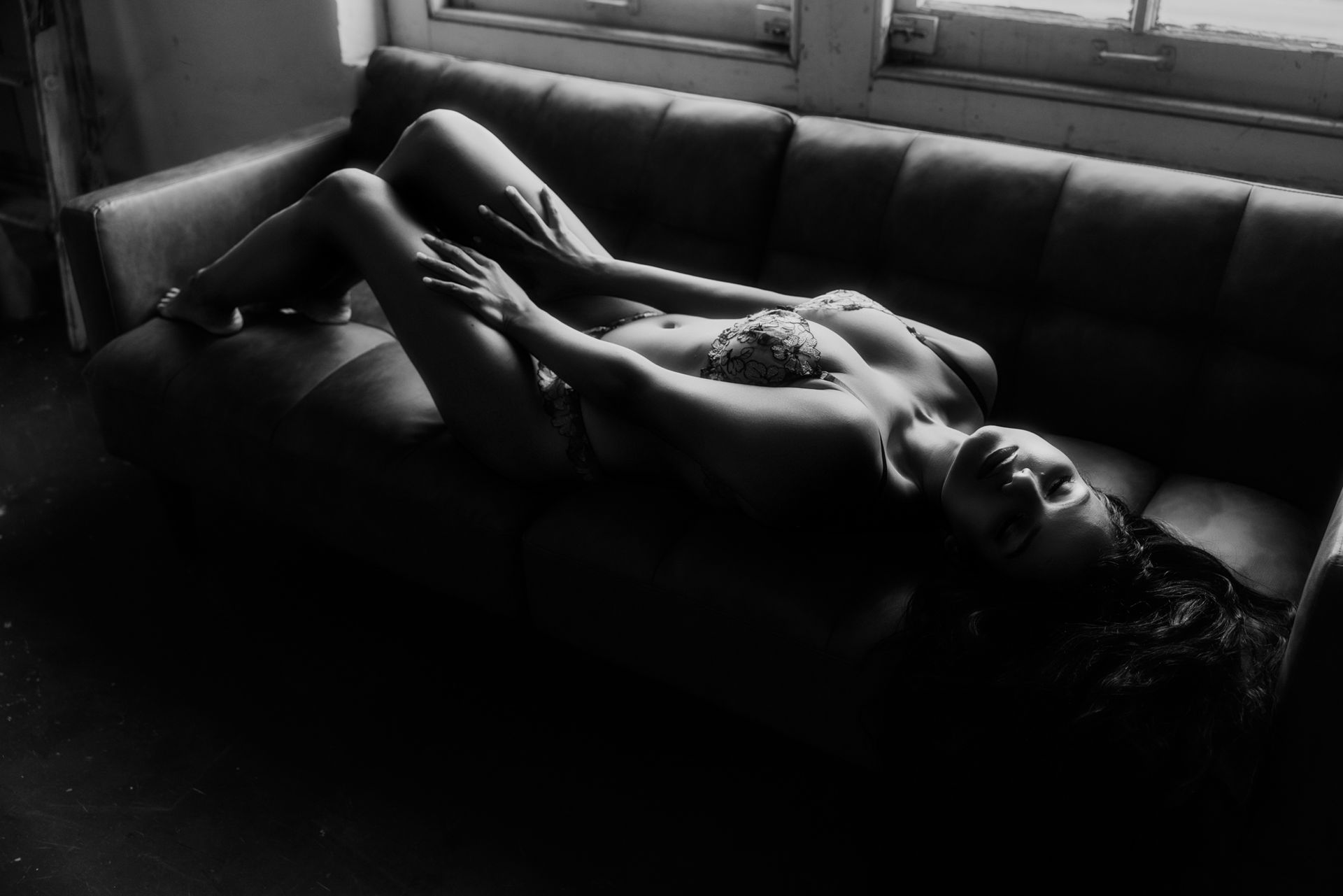 Woman reclining on a dark couch near a window. Black and white photo; she's partially nude, with arms crossed over her chest.
