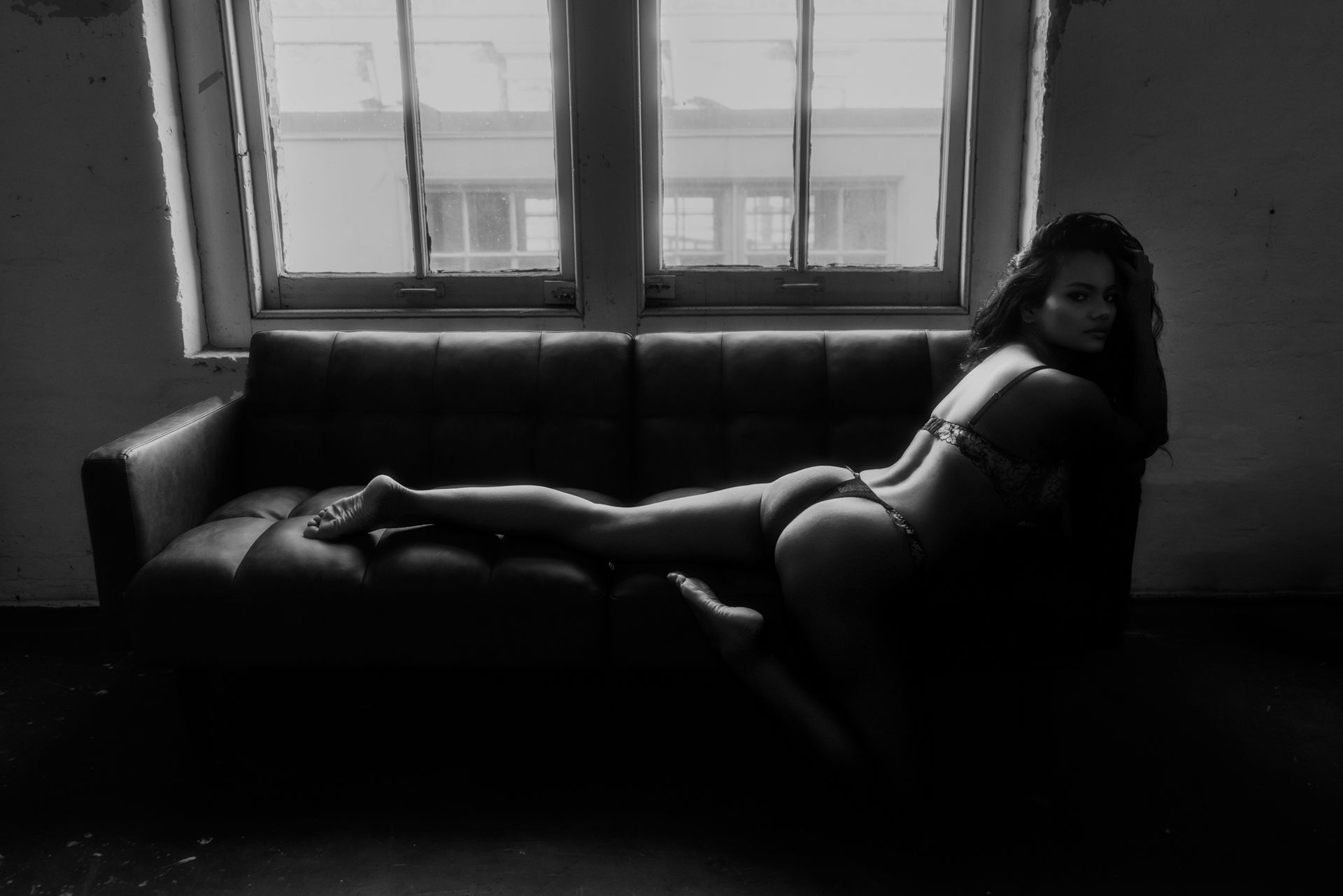 Woman reclining on a dark couch in a room, lit by sunlight from a window behind her.