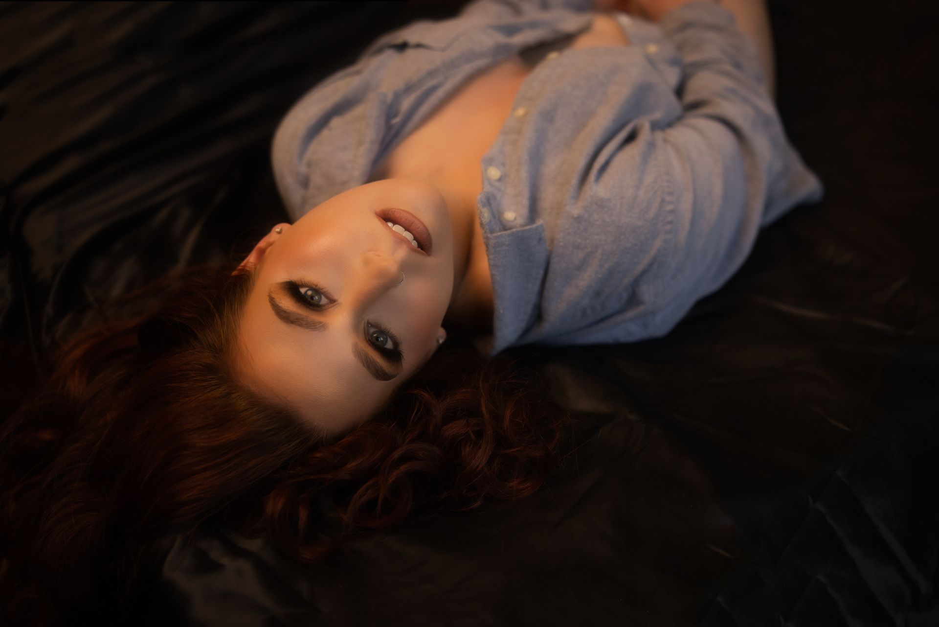 Woman with red curly hair, wearing an open blue shirt, reclines on a dark surface and smiles at the camera.