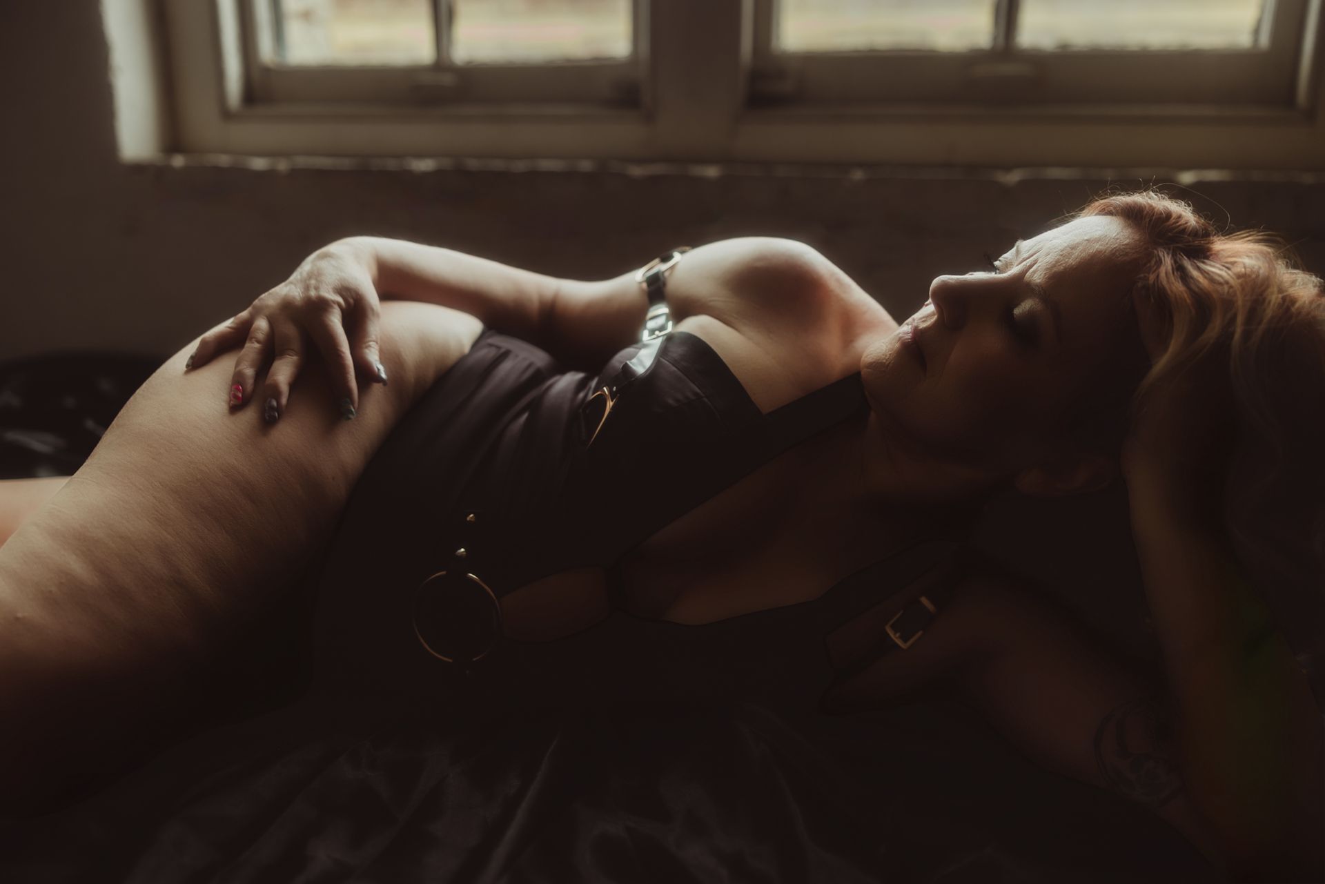 Woman reclines on a bed near a window, wearing a dark corset, hands raised, and hair styled.