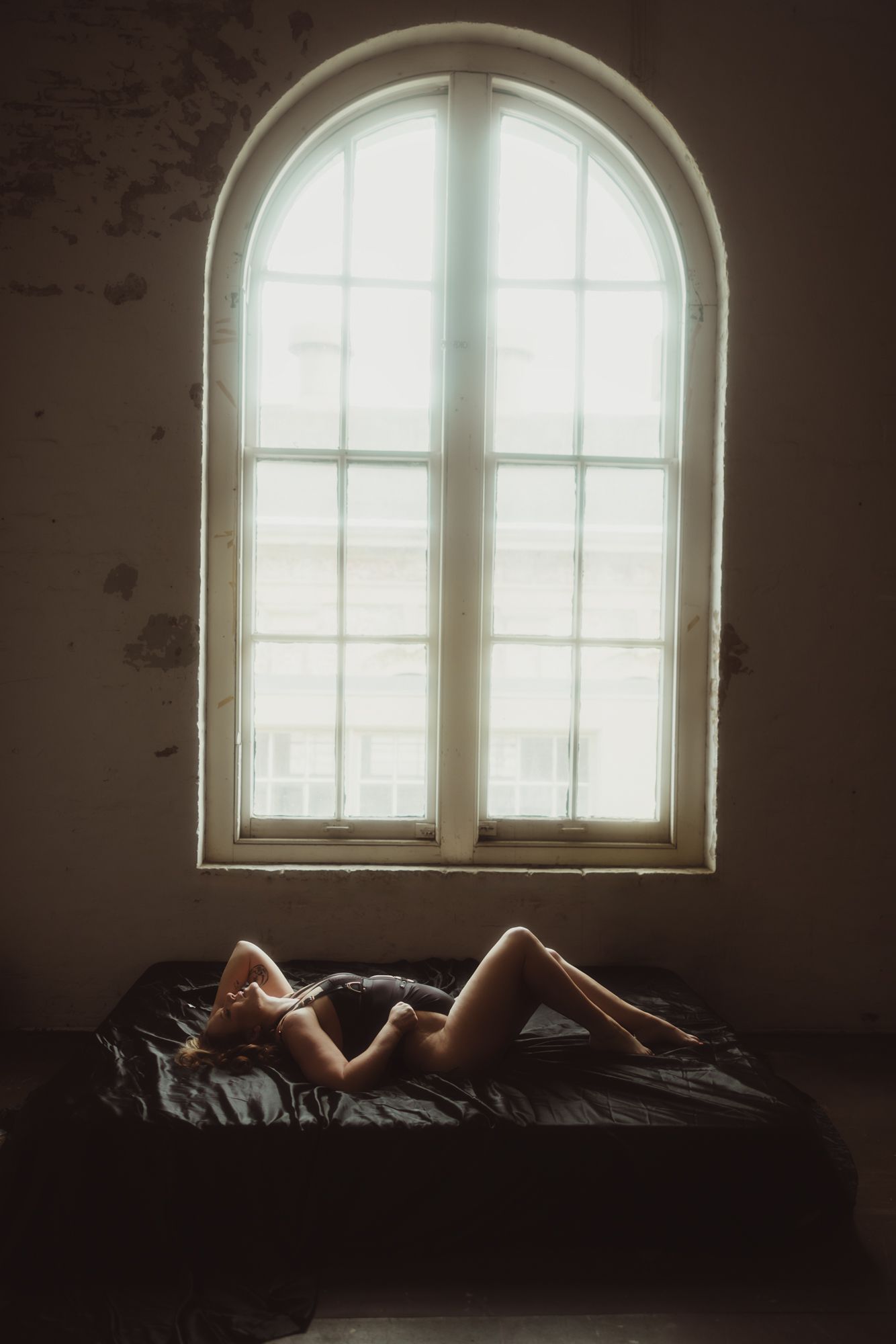 Woman lying on a bed in front of a large, arched window. The room is dimly lit.