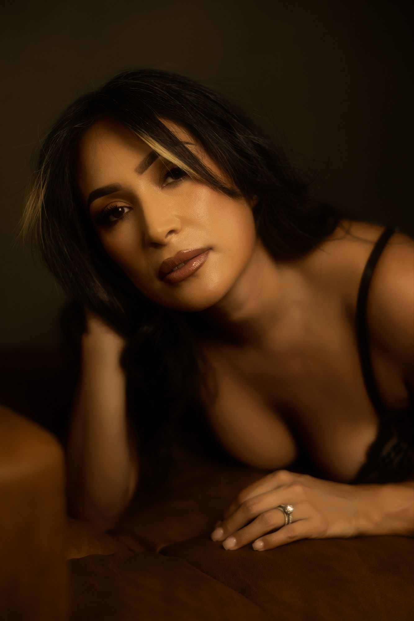 Woman with dark hair and a ring, laying on her arm, looking at the camera. Warm tones, dark background.