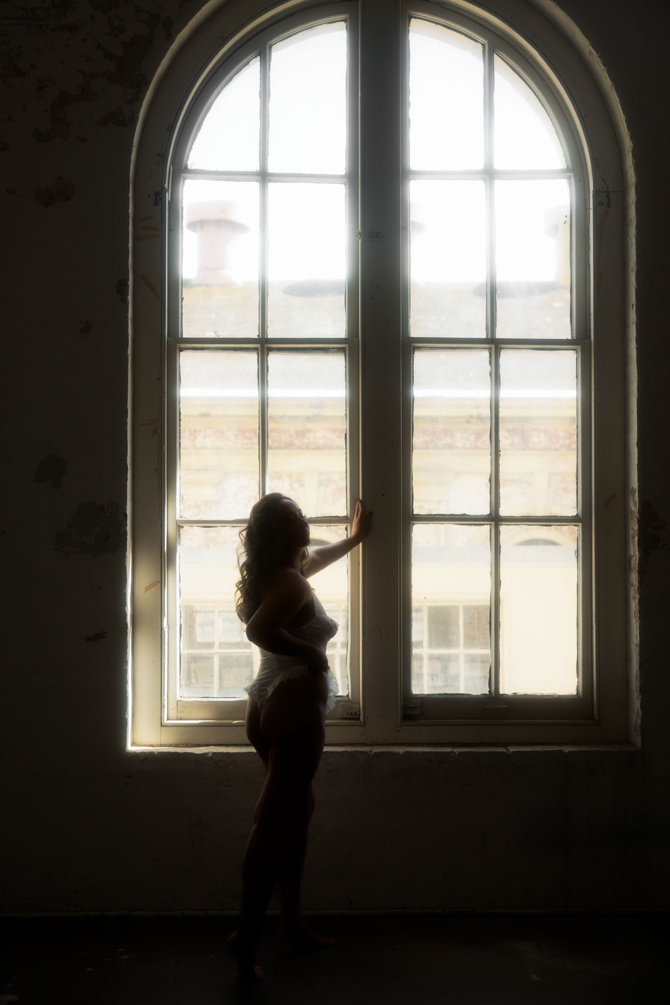 Woman in lingerie silhouetted by a large arched window. Light streams in, highlighting her figure, with a building visible outside.