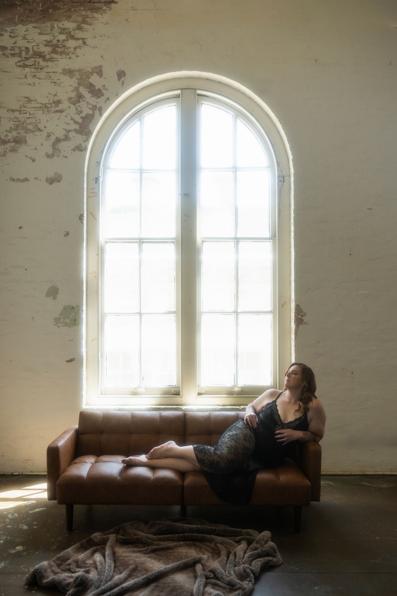 Woman in a black dress reclines on a brown leather couch in front of a large arched window. A faux fur blanket lies on the floor.