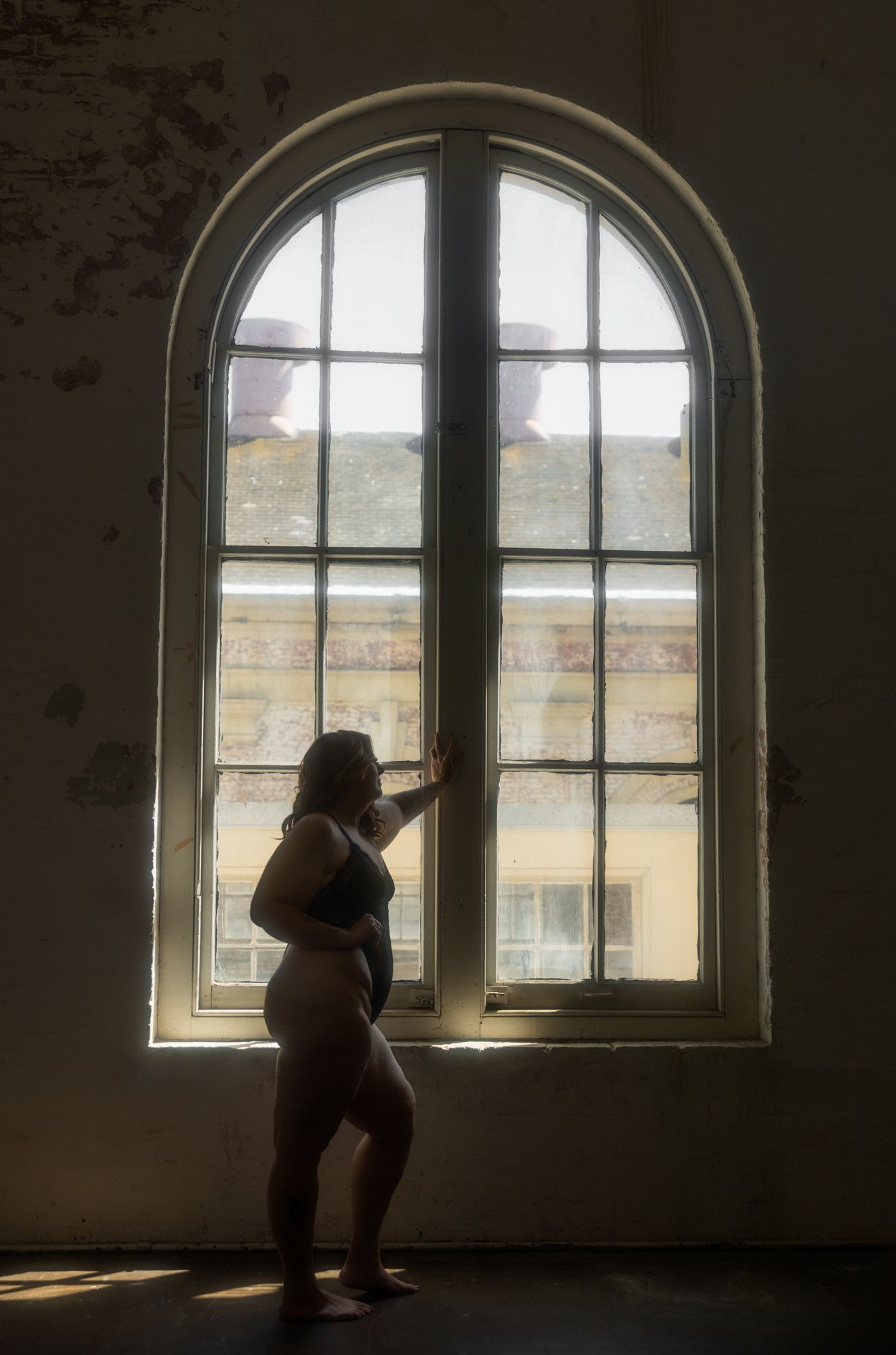 Woman in lingerie stands silhouetted by arched window, touching the frame. Sunlight streams in, highlighting the peeling wall.