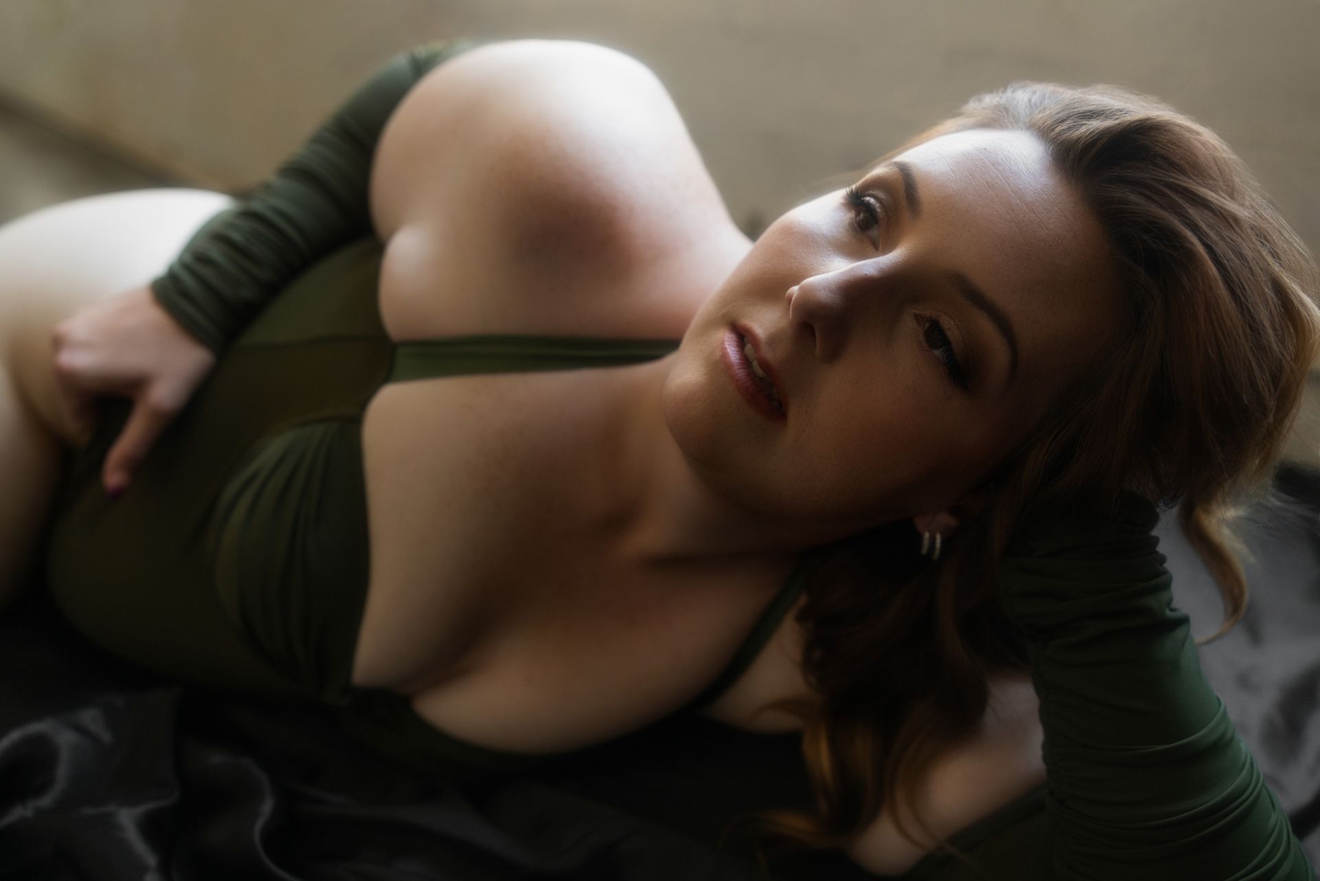 Woman with fair skin wearing an olive-green bodysuit poses on black fabric, leaning on her arm. She looks slightly upward, with a soft expression.