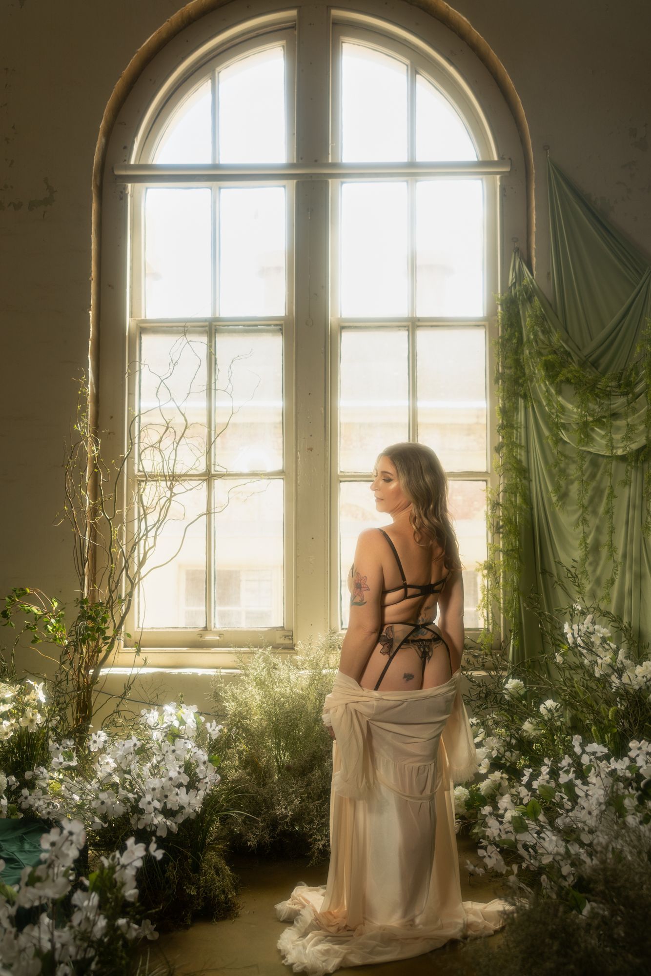 Woman in lingerie, facing away, stands near a large window in a room filled with plants and flowers.