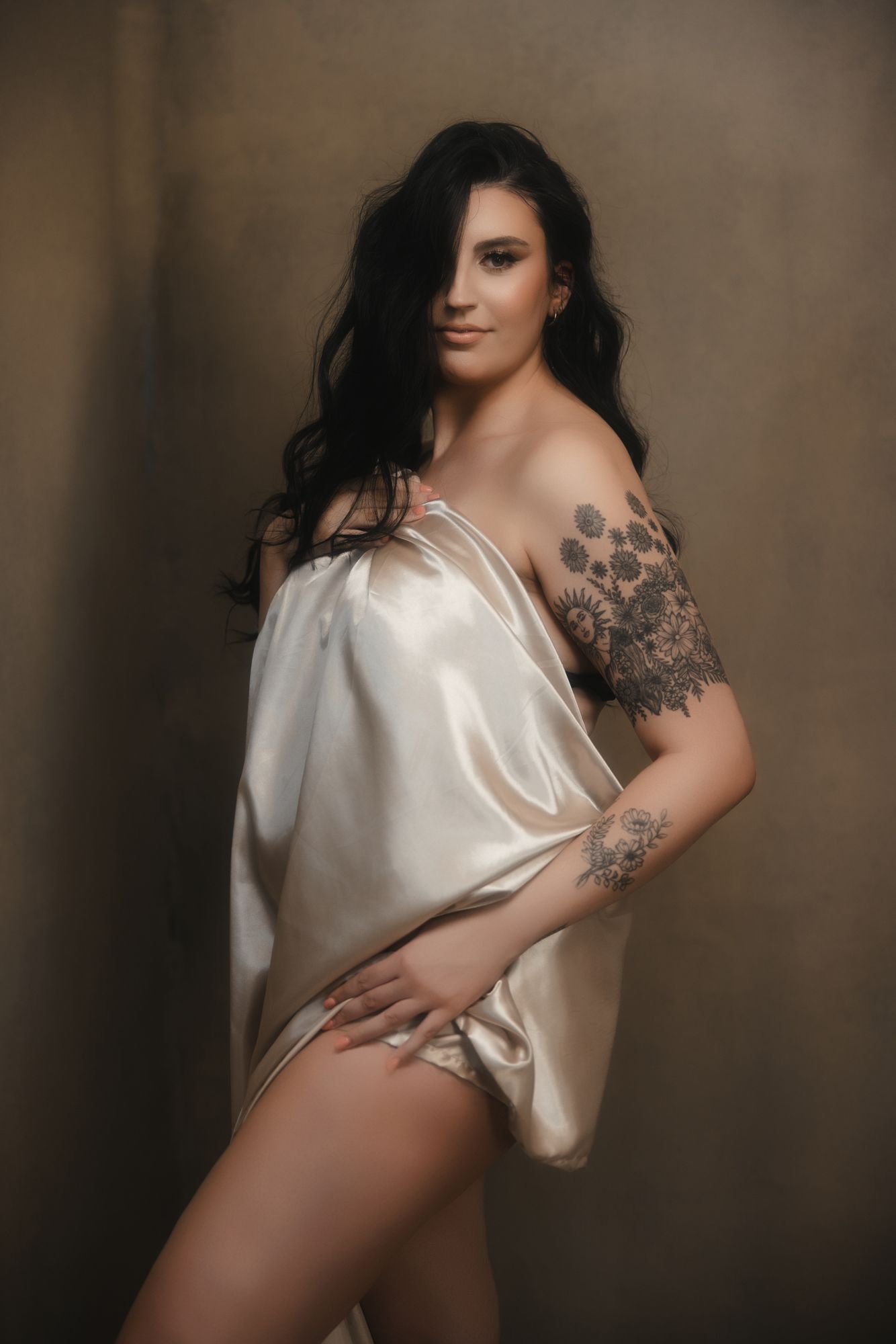Woman with long dark hair, draped in a cream-colored fabric, poses with a hand on her hip, tattoos visible on her arm.