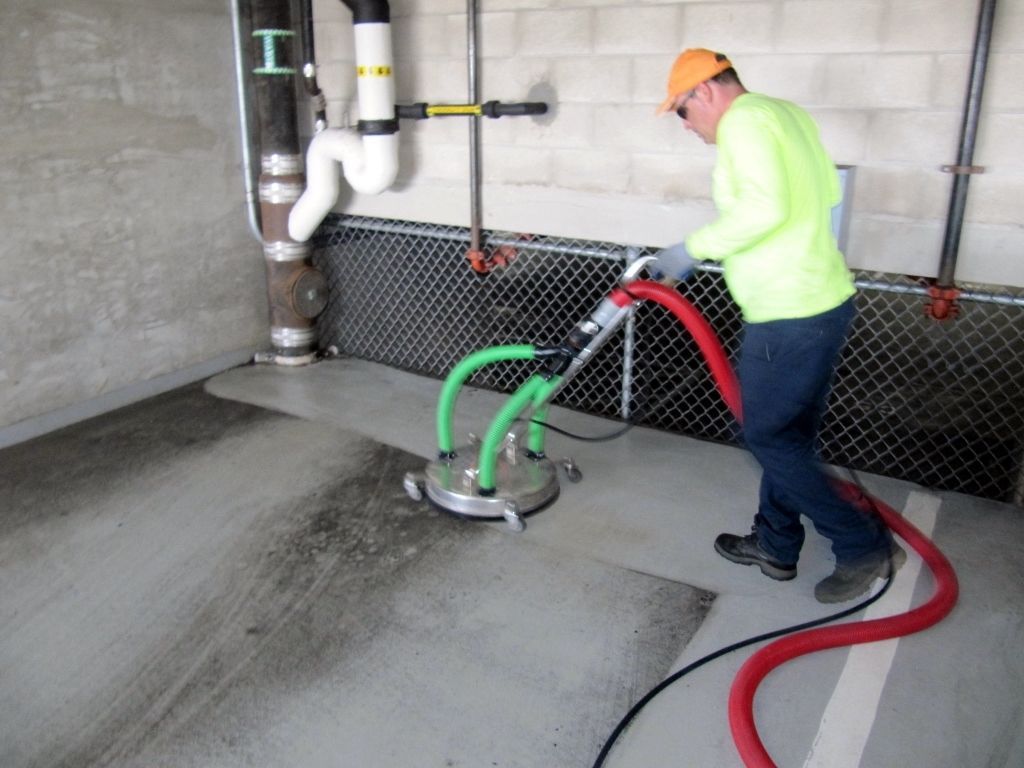 A worker in a bright yellow shirt uses a floor cleaning machine to wash a concrete garage floor.