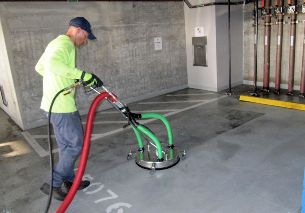A worker in a high-visibility lime shirt uses a surface cleaner to power wash a grey concrete parking garage floor.