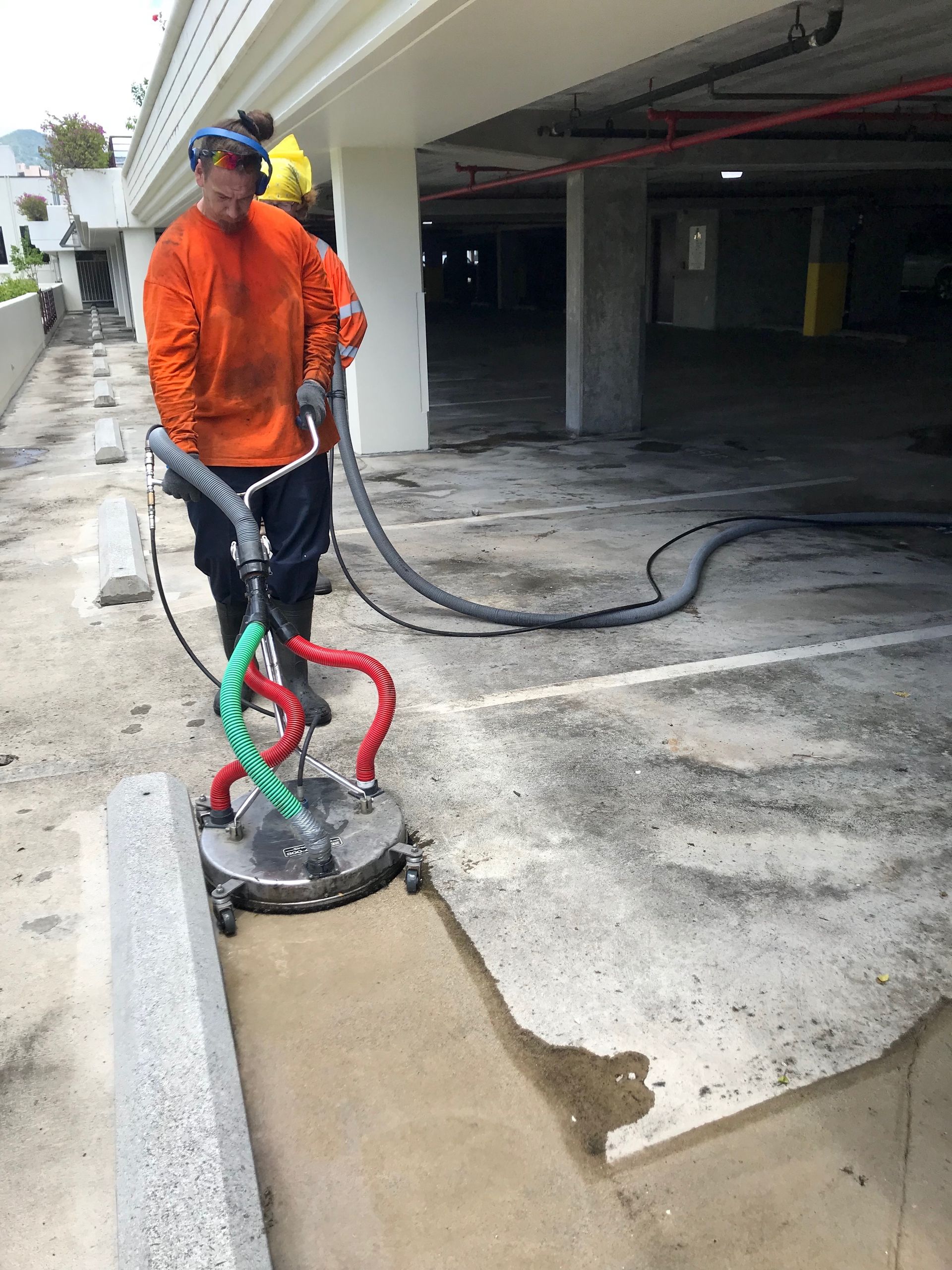 A person in orange work clothing uses a circular pressure washer to clean a concrete parking garage floor.