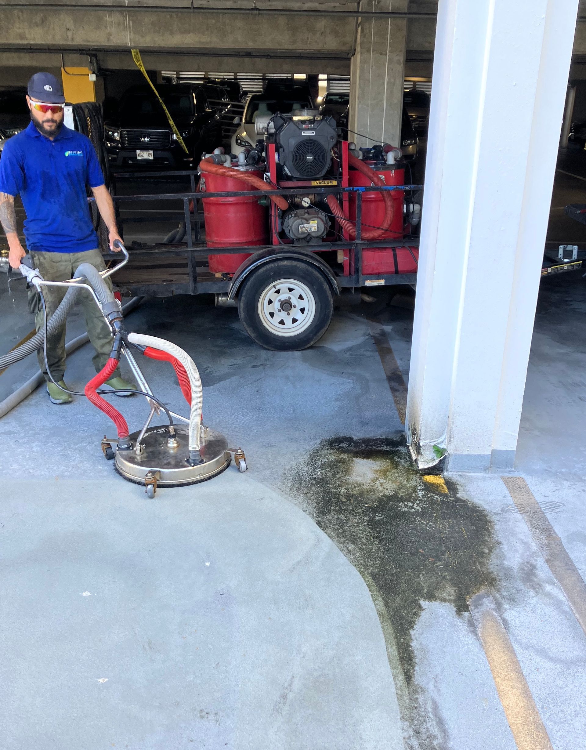 A worker in a parking garage uses a surface cleaning tool next to a trailer-mounted engine and a dirty, wet floor area.