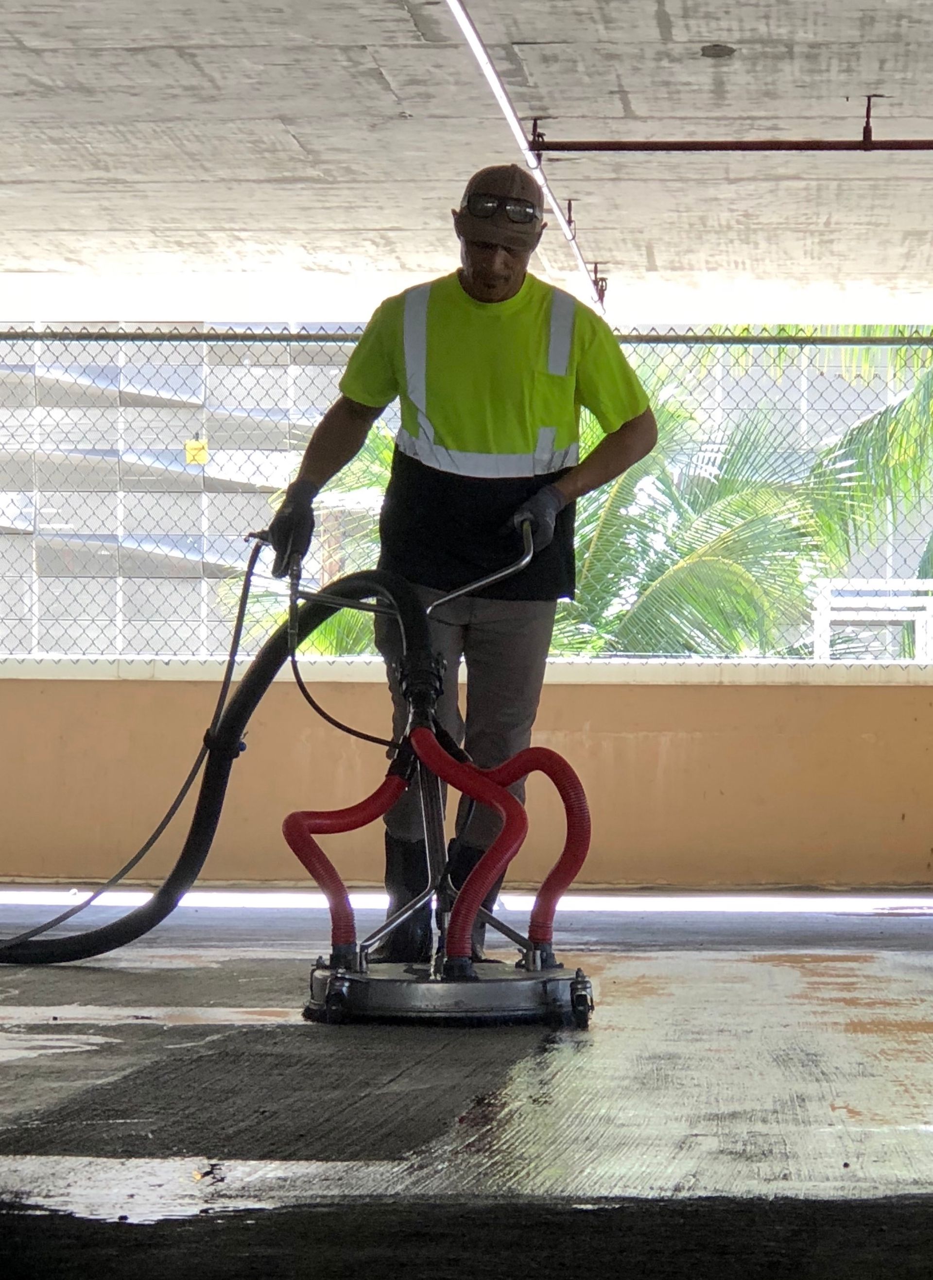 A worker in a high-visibility shirt uses a floor surface cleaner machine on a concrete parking garage deck.