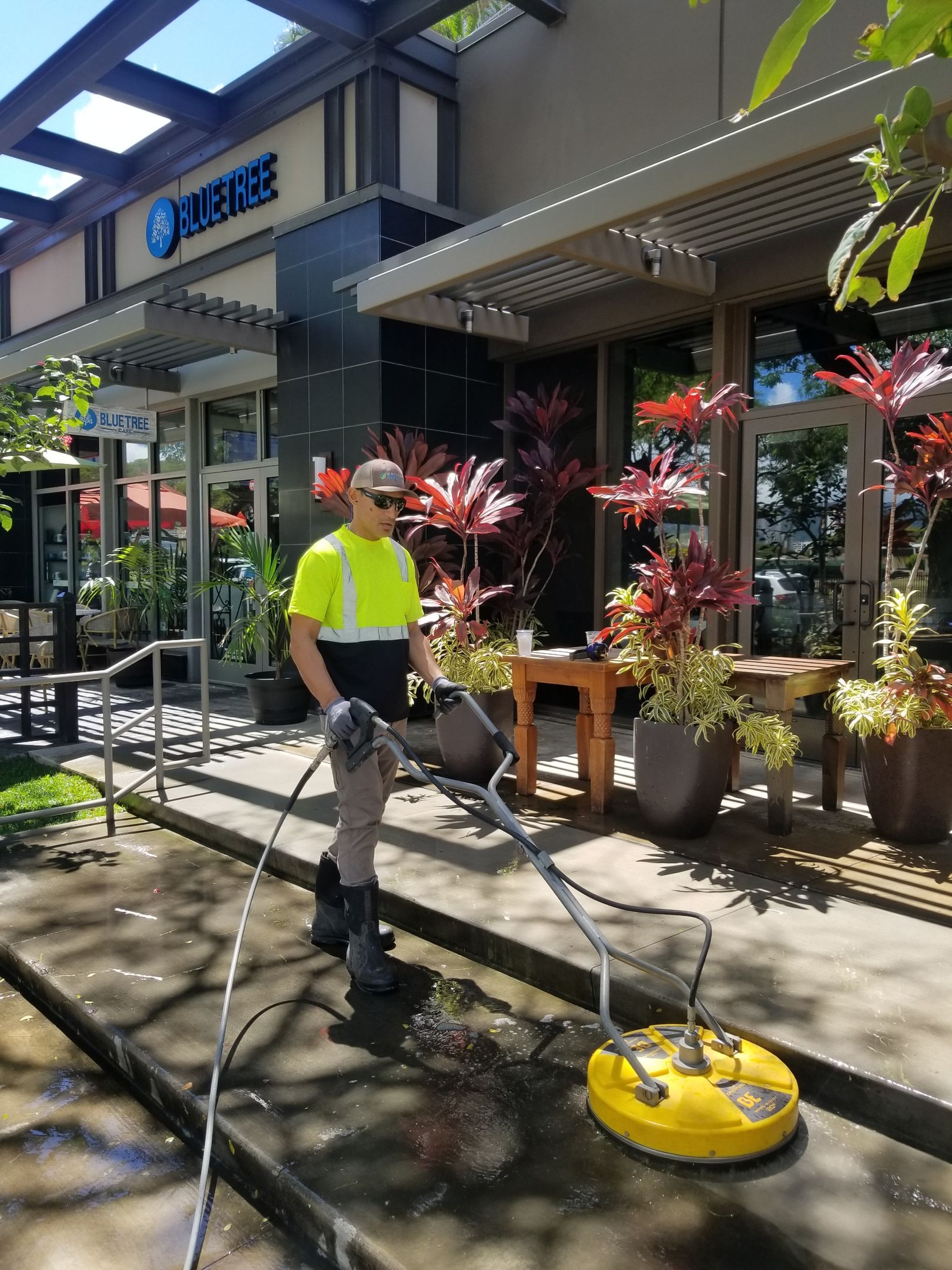 Worker using a yellow floor scrubber outside a storefront under a shaded walkway.