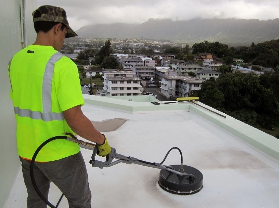 A person in a high-visibility vest uses a pressure washer surface cleaner on a flat white roof overlooking a town.