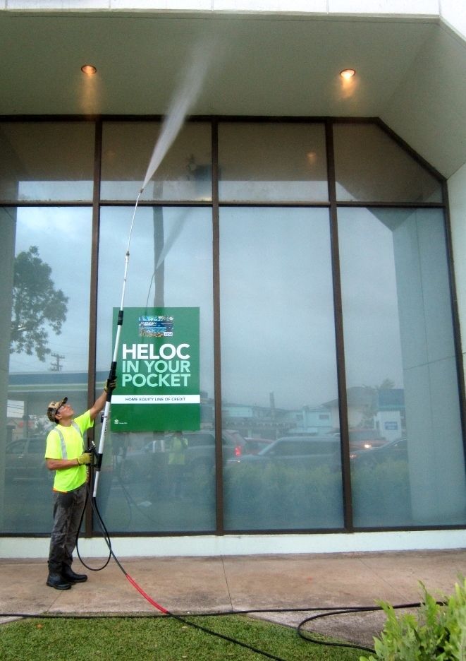 Worker in neon vest pressure-washes a glass storefront with a green sign.