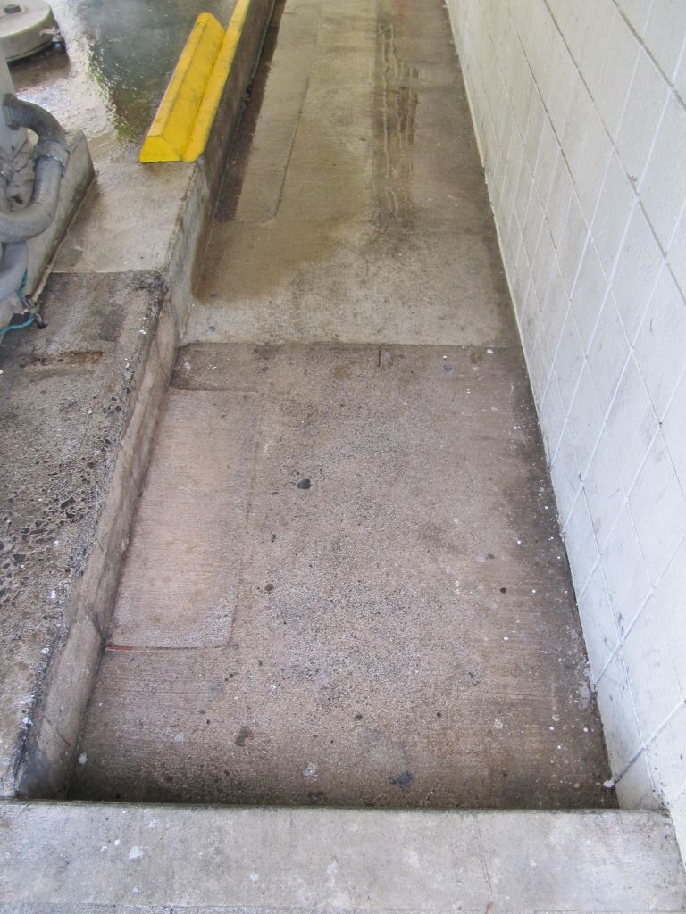 Narrow concrete ramp beside a white wall with a yellow curb stop and gravel along the edge