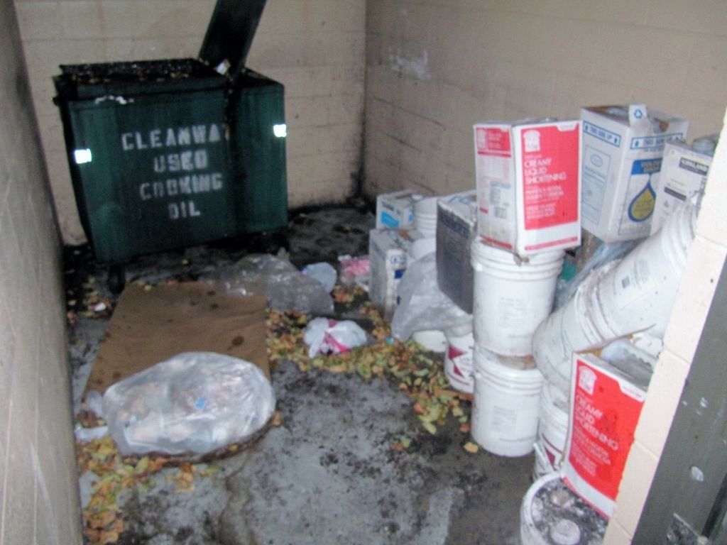 Trash and paint cans scattered in a dirty corner by a wall, with a green dumpster and debris on the floor.