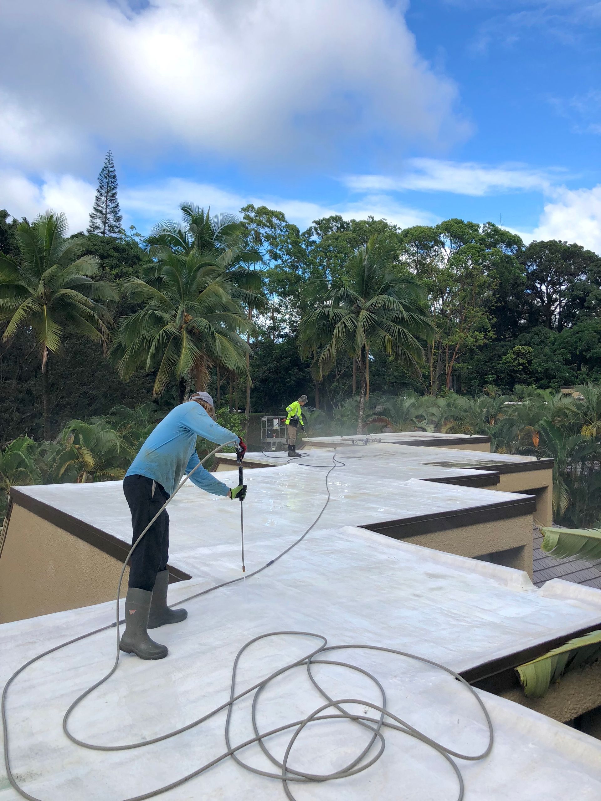 A person in a blue long-sleeved shirt and boots power washes a flat roof with trees in the background.