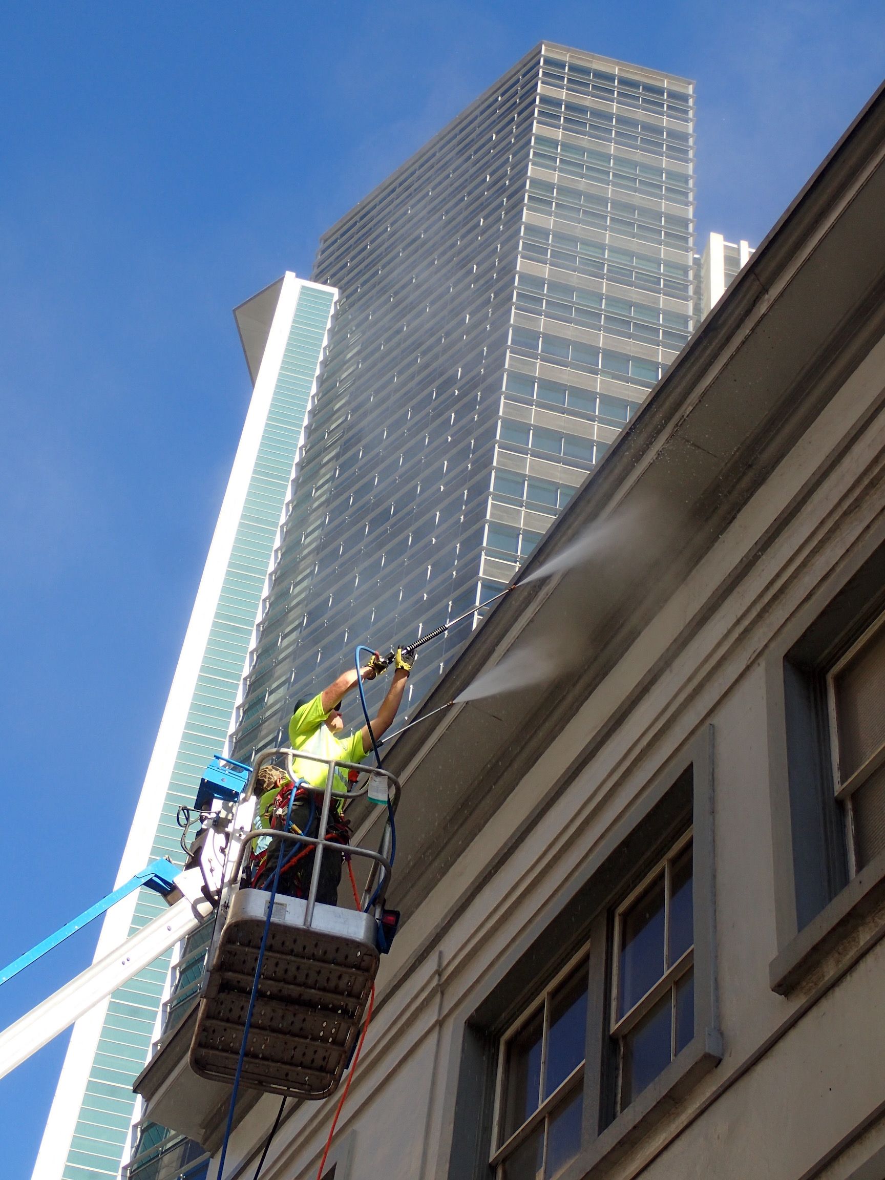 A person in a high-visibility vest uses a pressure washer to clean a building ledge from a hydraulic lift.