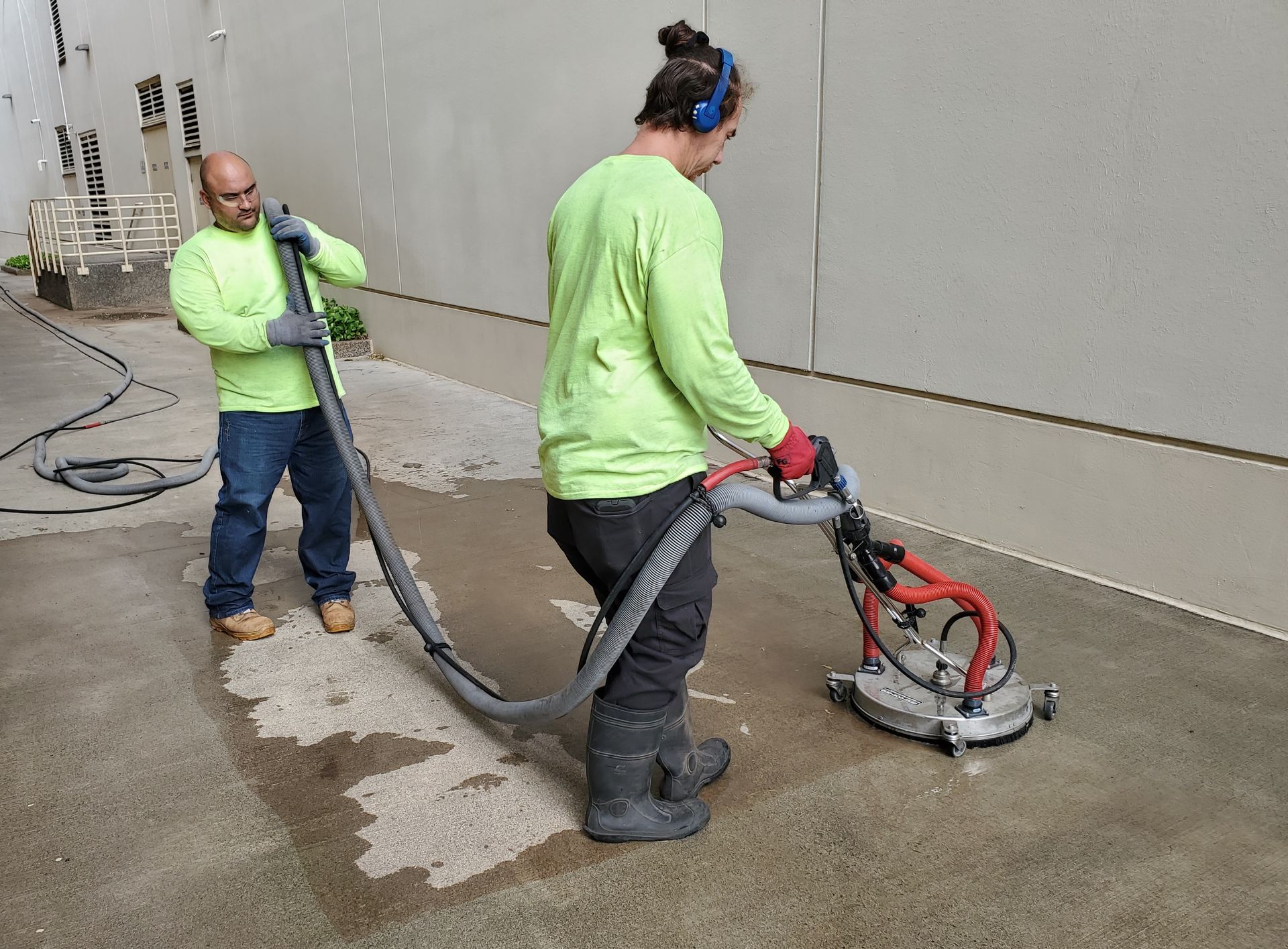 Two workers using a floor grinder on concrete, one holding a vacuum hose to control dust.