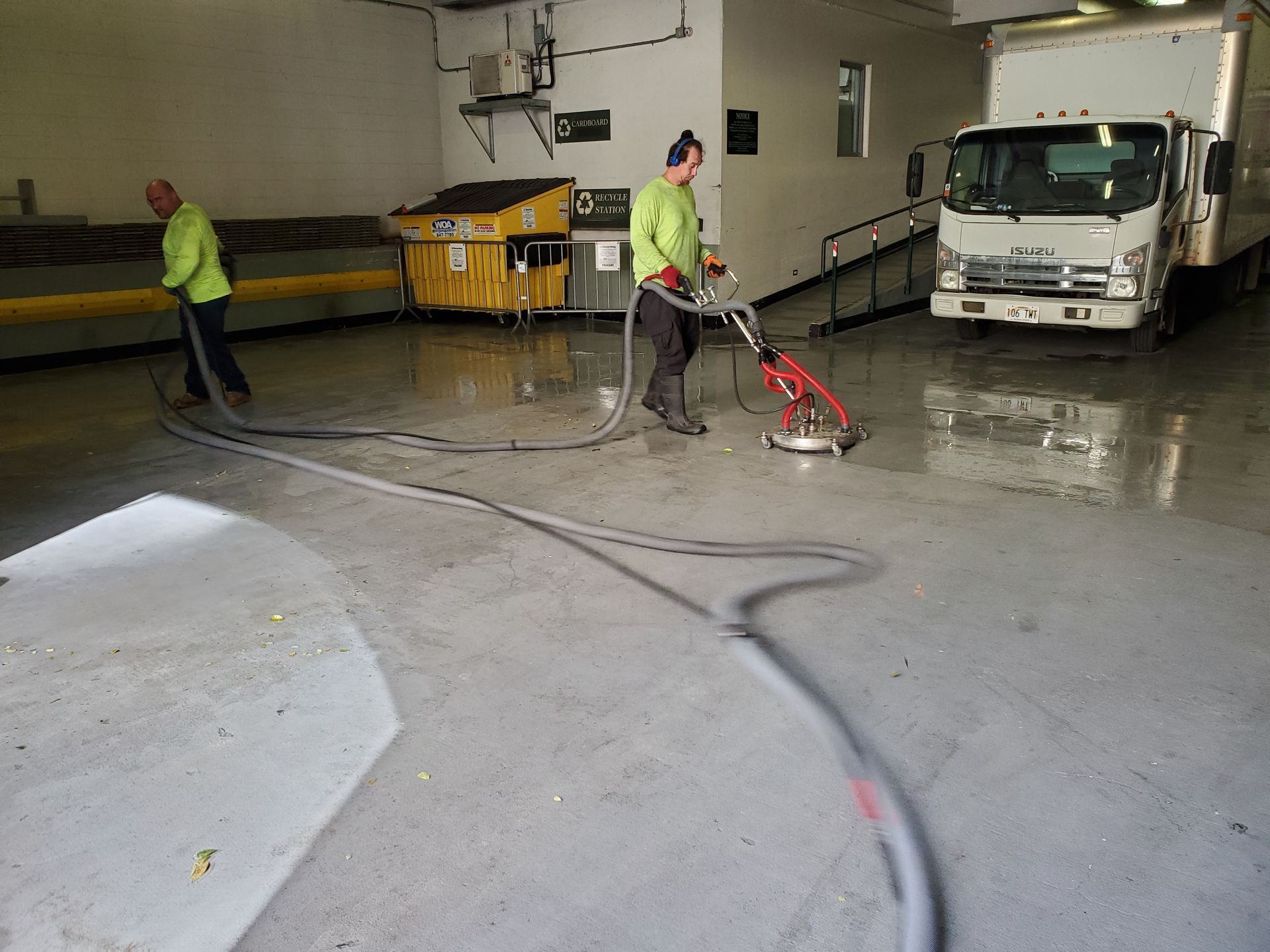 Workers pressure-washing a wet warehouse floor beside a truck and yellow bins