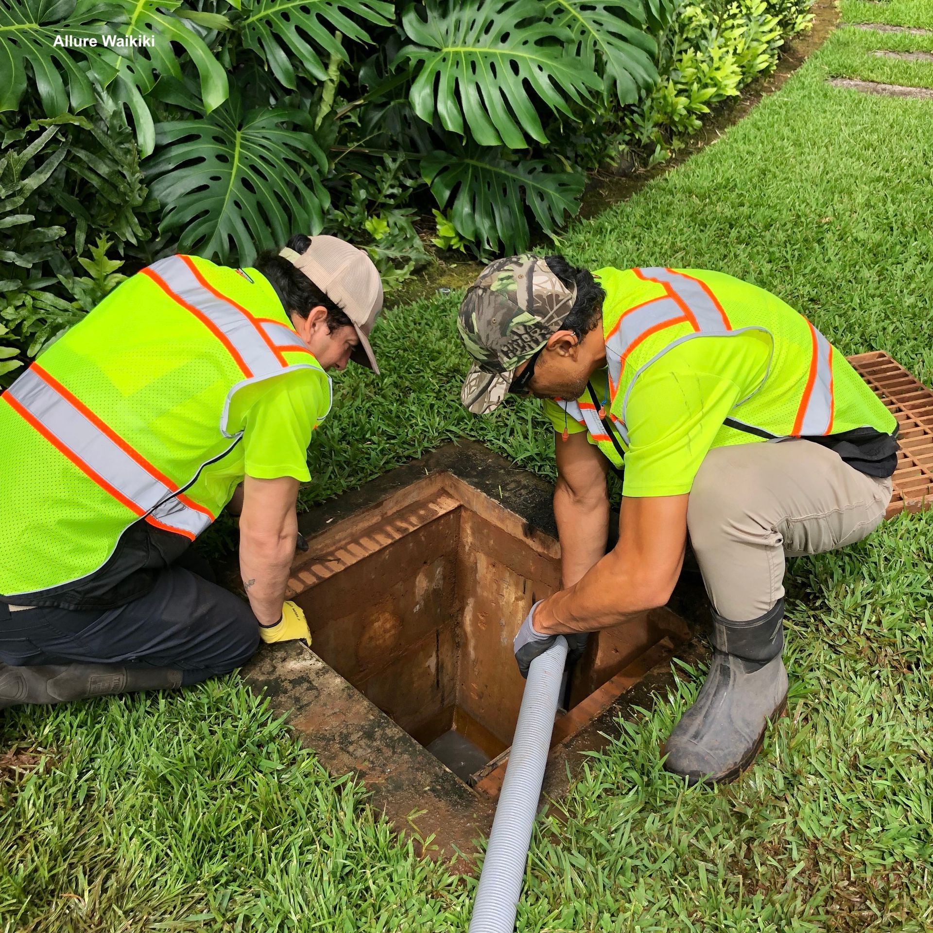 Two workers in neon safety vests and hats crouch by an open utility hole, using a hose for maintenance work on a lawn.