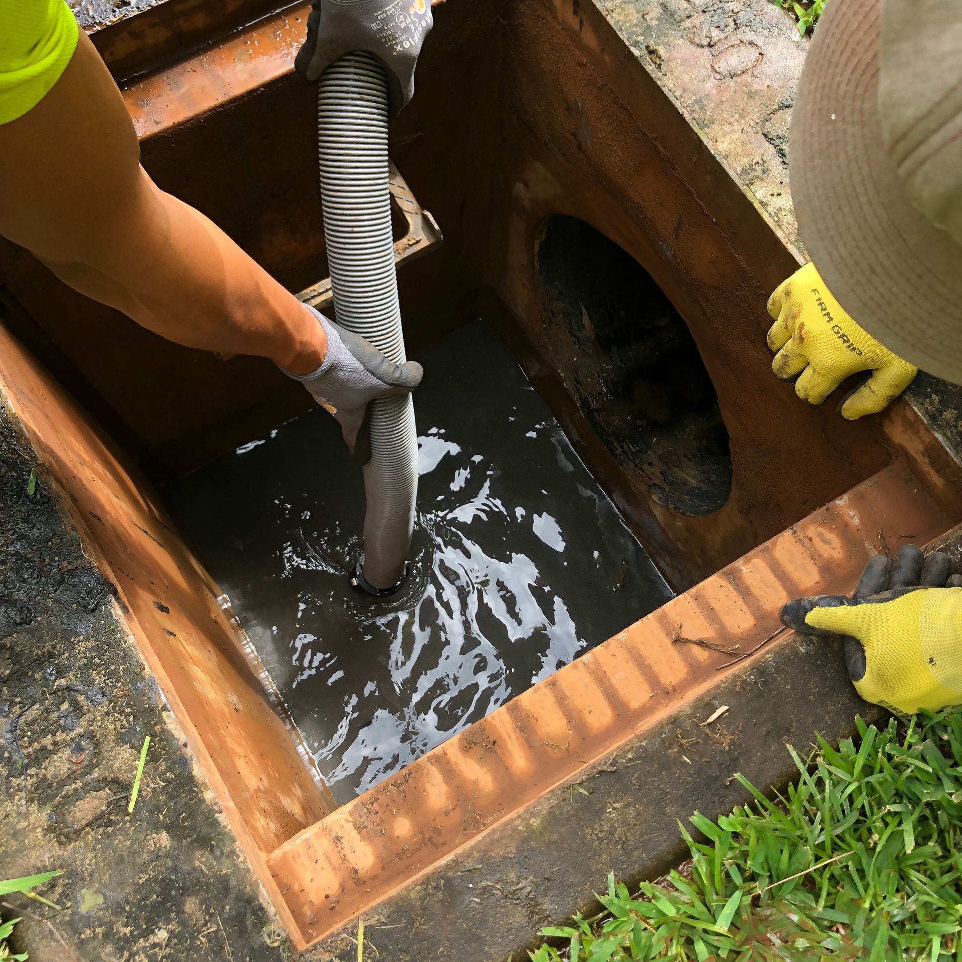 Workers in gloves use a suction hose to clear murky water from a concrete storm drain box.