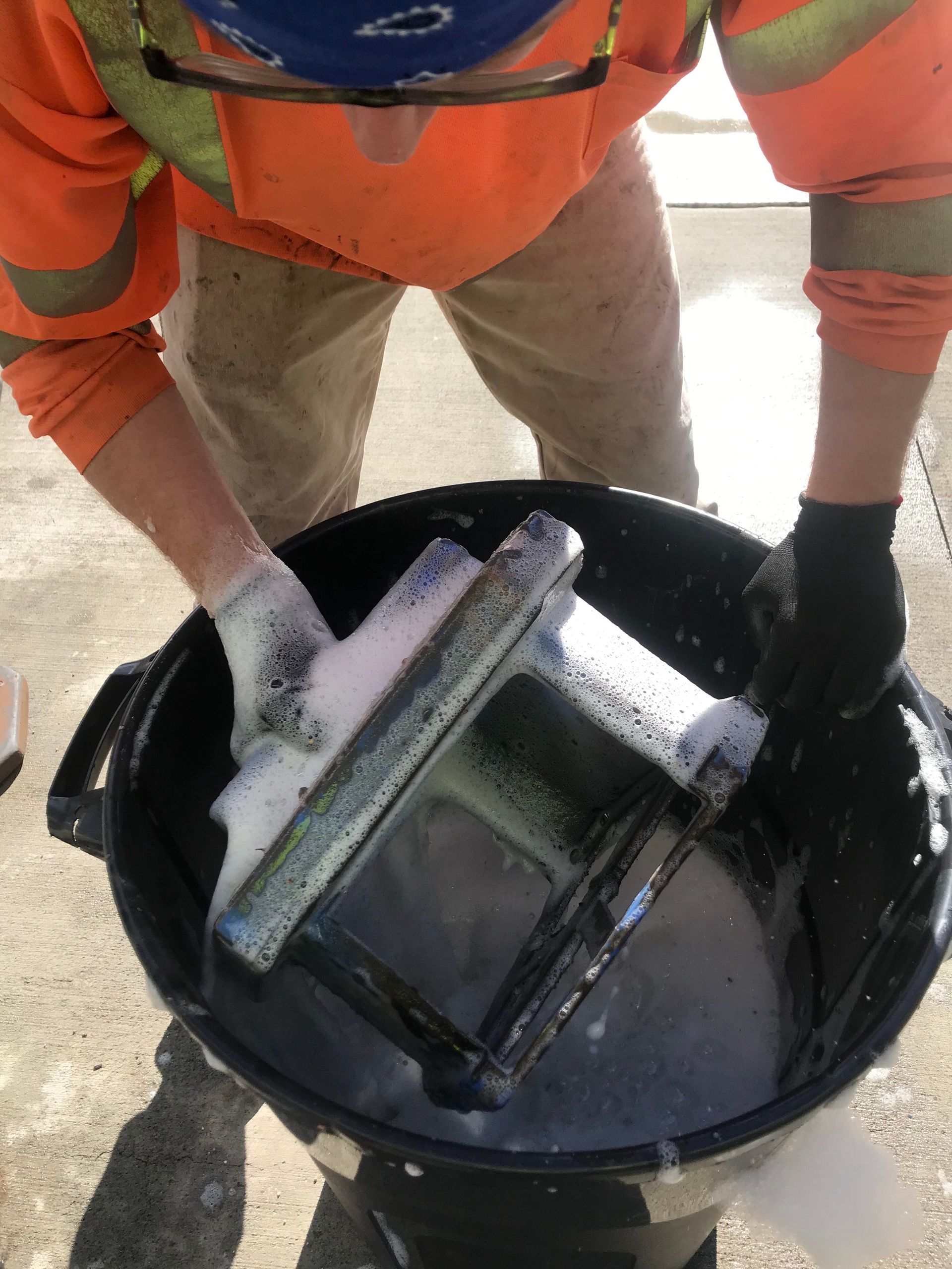 Worker in orange safety vest handling tools in a black bucket outdoors.
