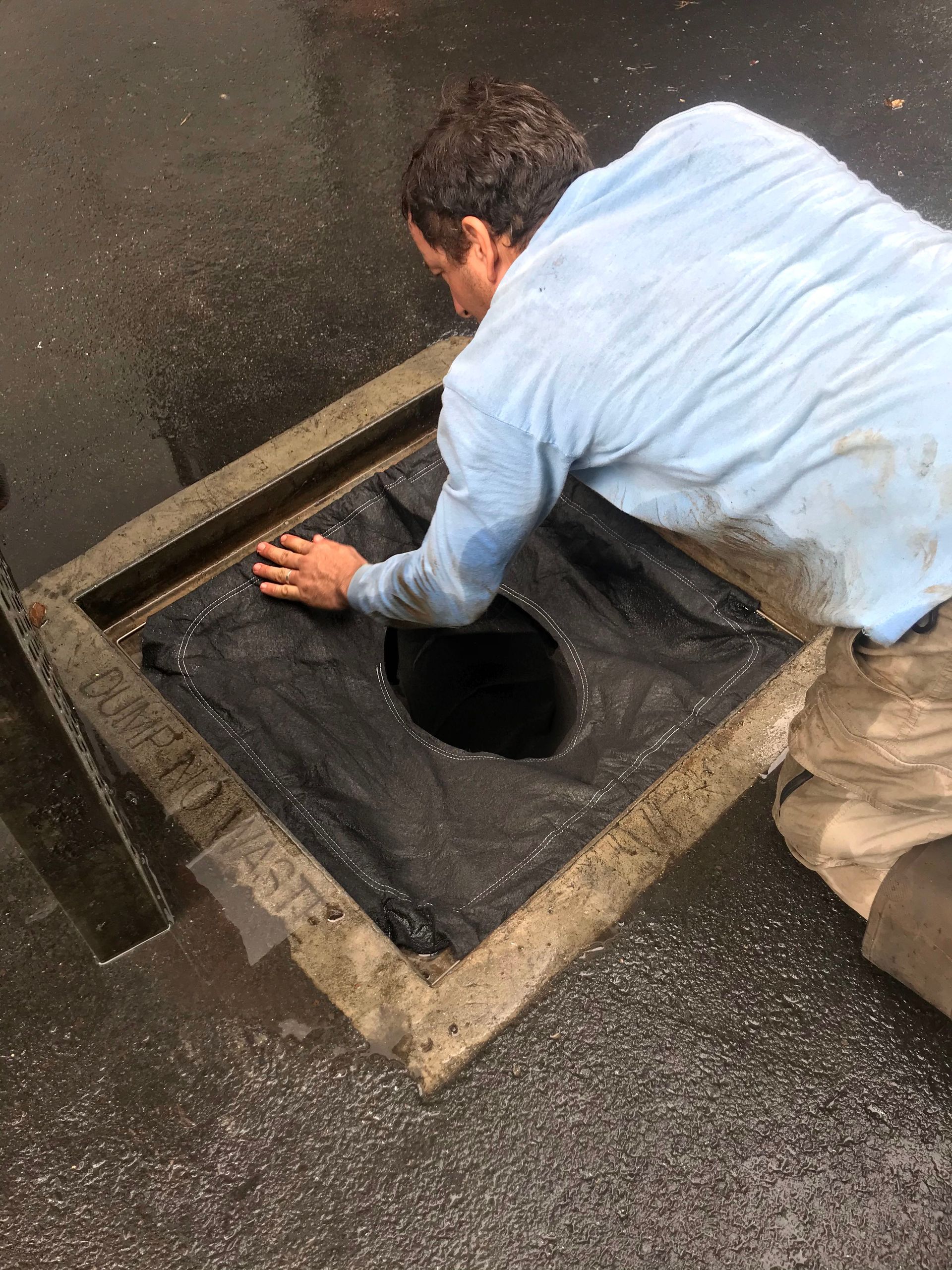 Worker crouching over a floor drain or opening, reaching into a concrete hatch in a wet industrial area.