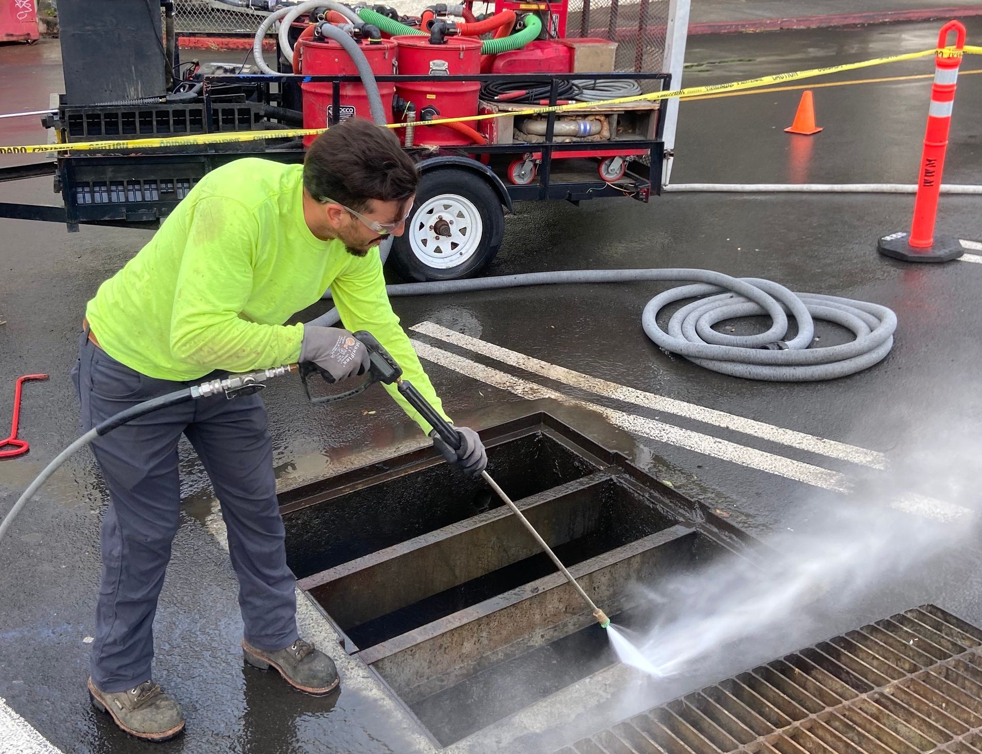 Worker in neon shirt pressure-washes an open street drain beside a utility truck and traffic cones.