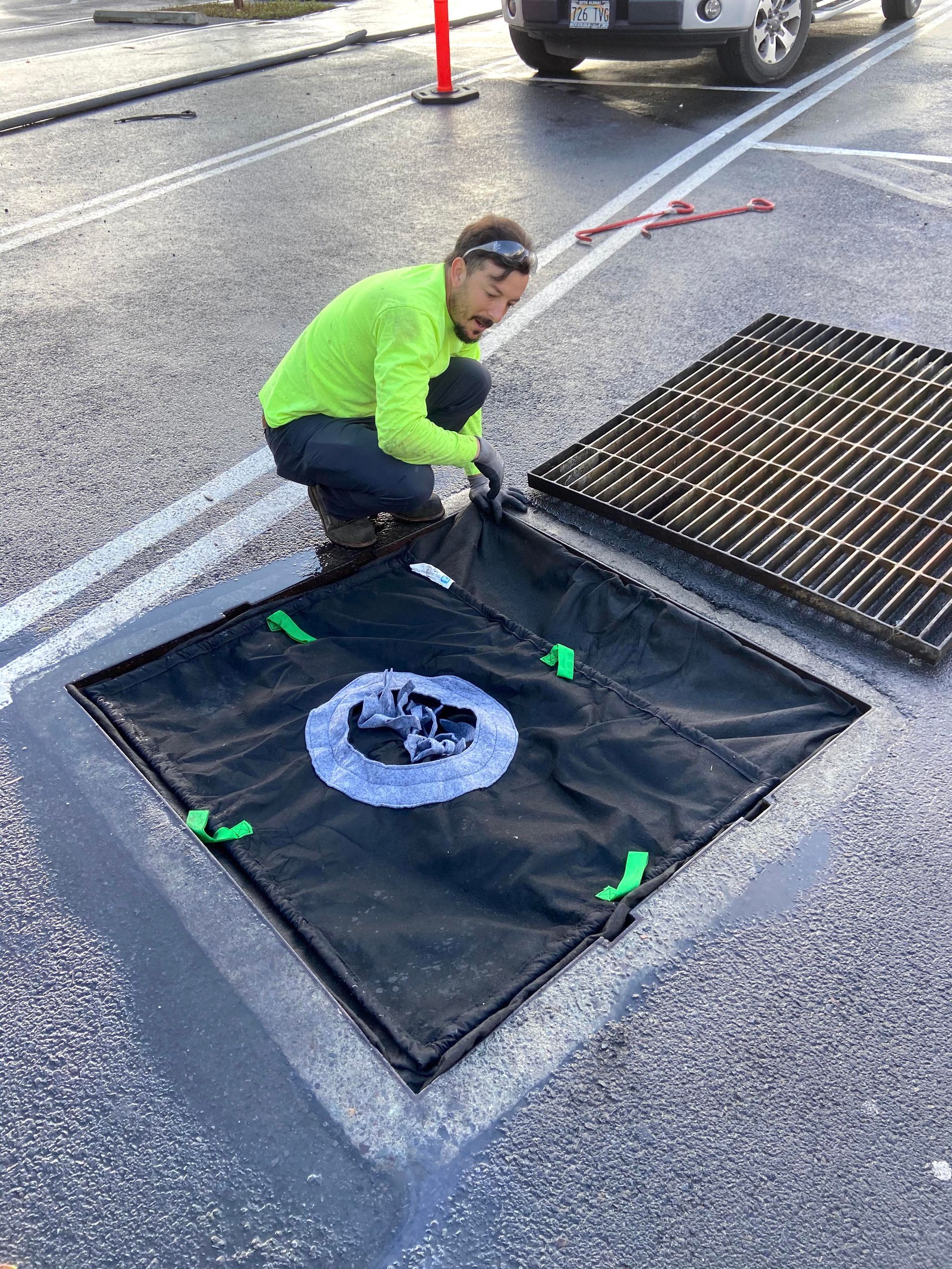 Worker in neon shirt kneeling beside an open storm drain on wet pavement, using a grate cover.