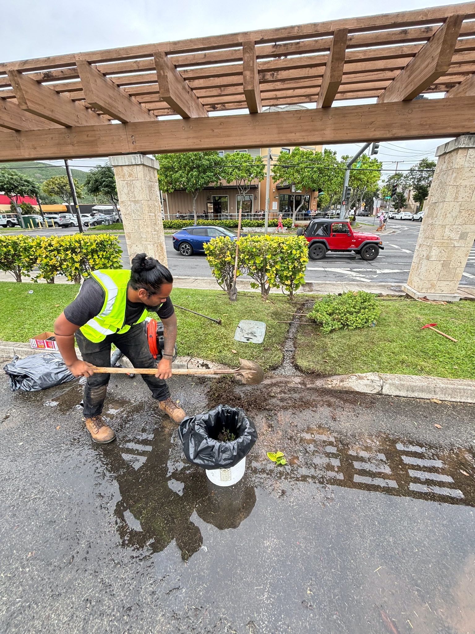Worker in a neon vest cleaning a puddle beside a landscaped walkway and wooden pergola.