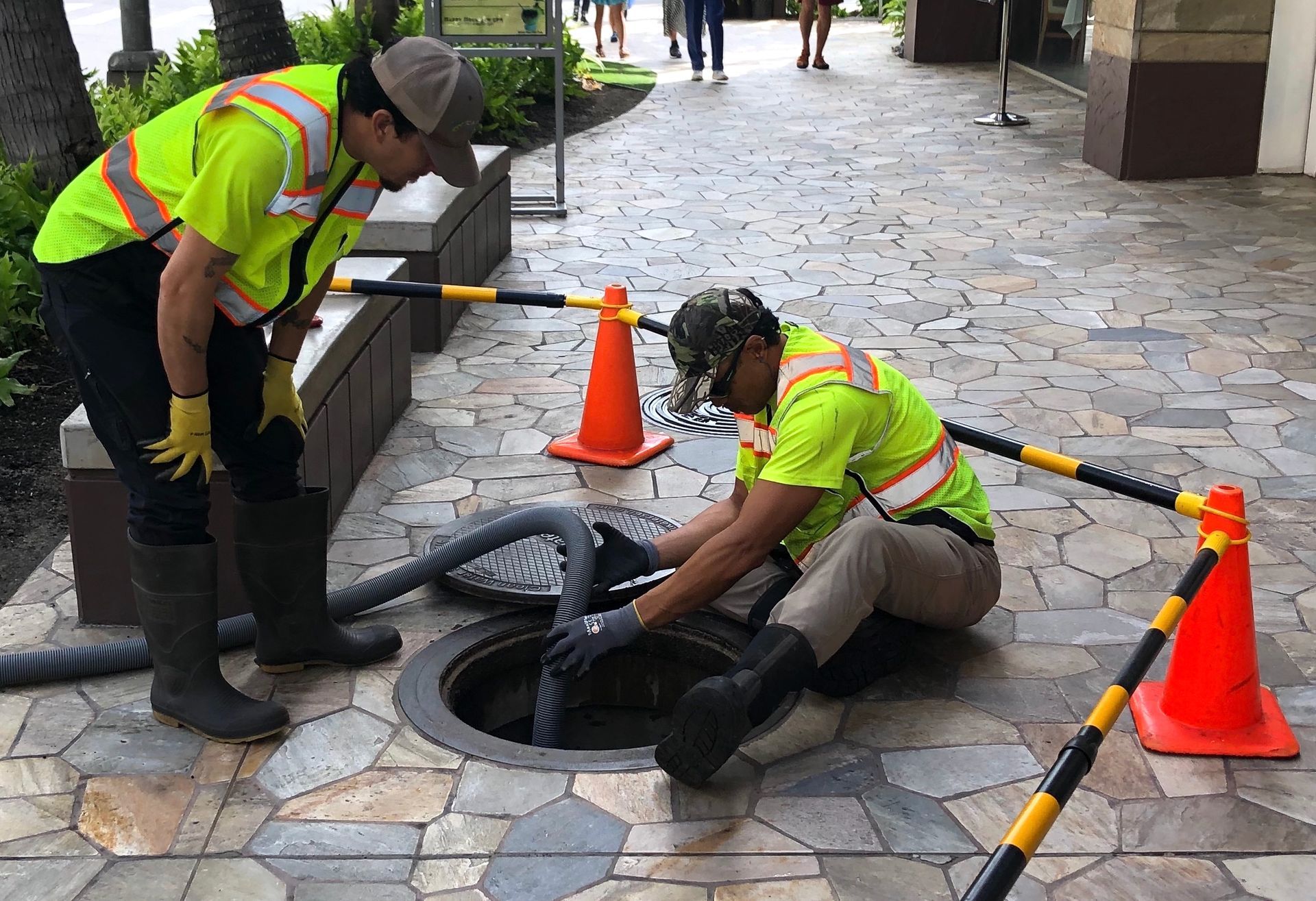 Two workers in neon vests and boots service an open sewer manhole on a stone-paved walkway marked by orange cones.