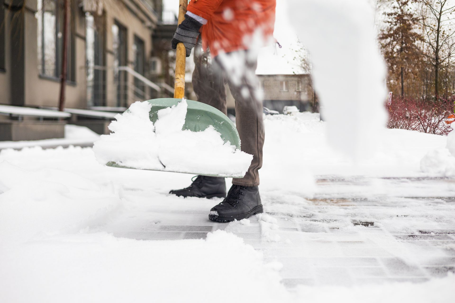 Person shoveling snow from a walkway with green shovel, orange jacket, winter boots, in front of a building.