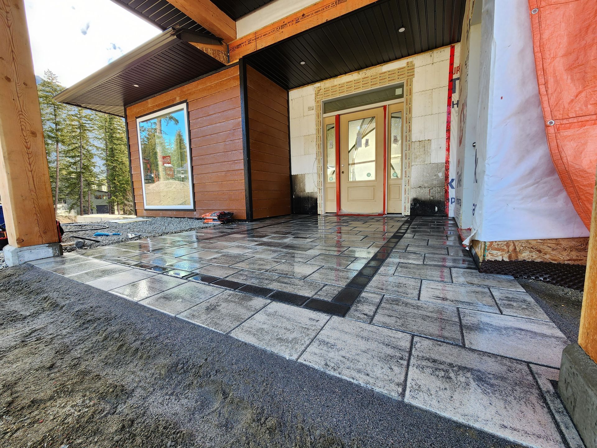 A newly paved stone patio with a dark border leads to a wooden front entrance under construction.