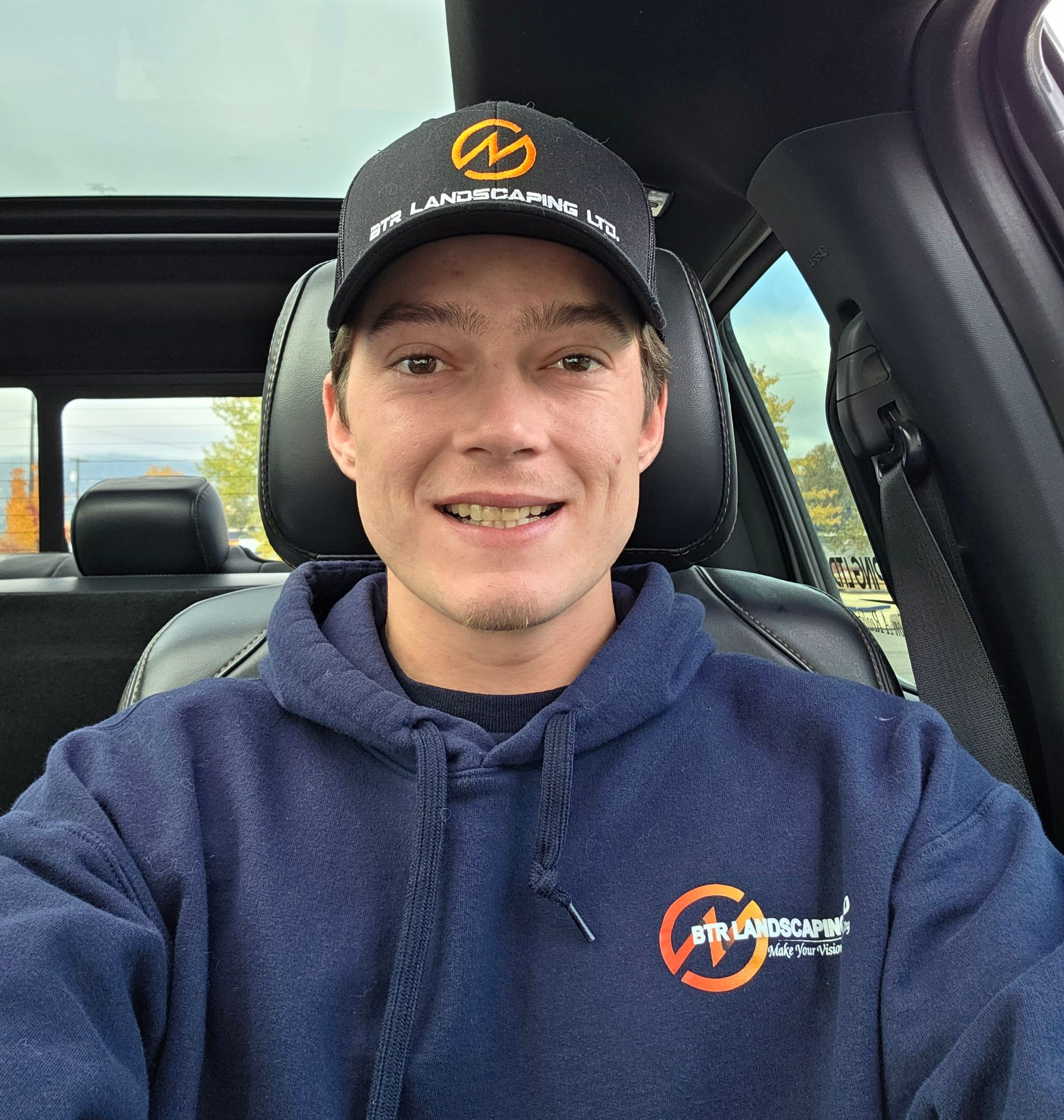 Person in a car wearing a navy hoodie and hat with a logo, smiling at the camera.