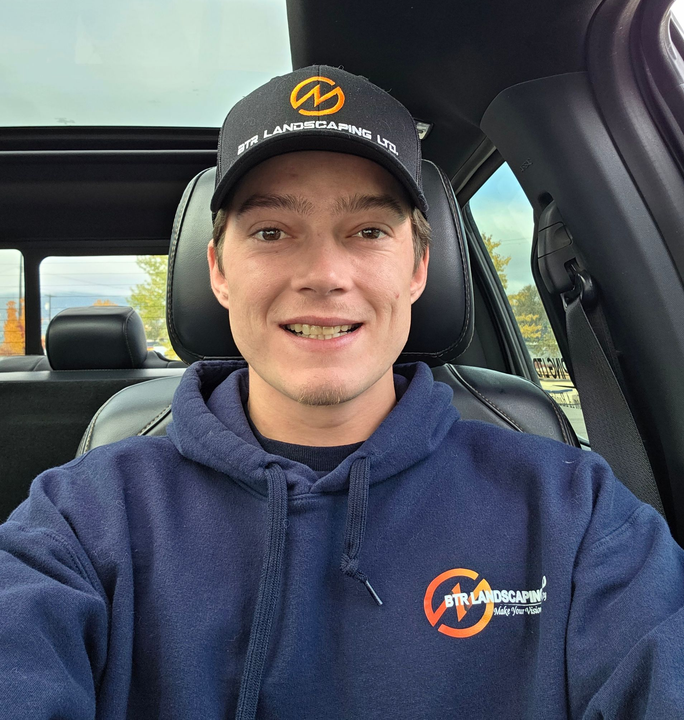 Person in a car wearing a navy hoodie and hat with a logo, smiling at the camera.