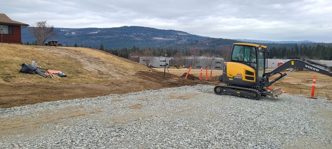 A yellow excavator on a gravel road with a grassy hill, house, and mountains in the background under a cloudy sky.