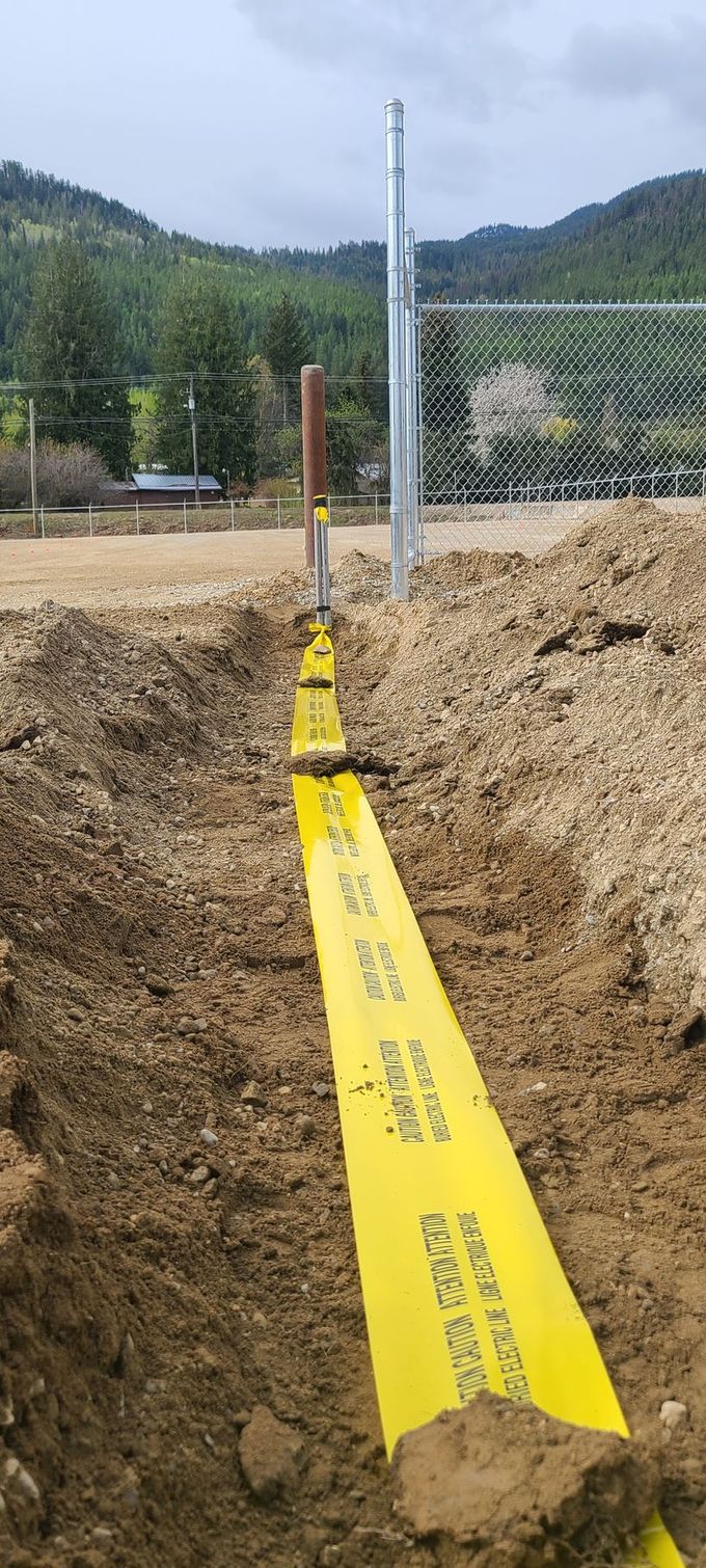 A trench with a yellow warning tape is visible, indicating buried utilities. A metal pole, likely a fence post, is in the background.