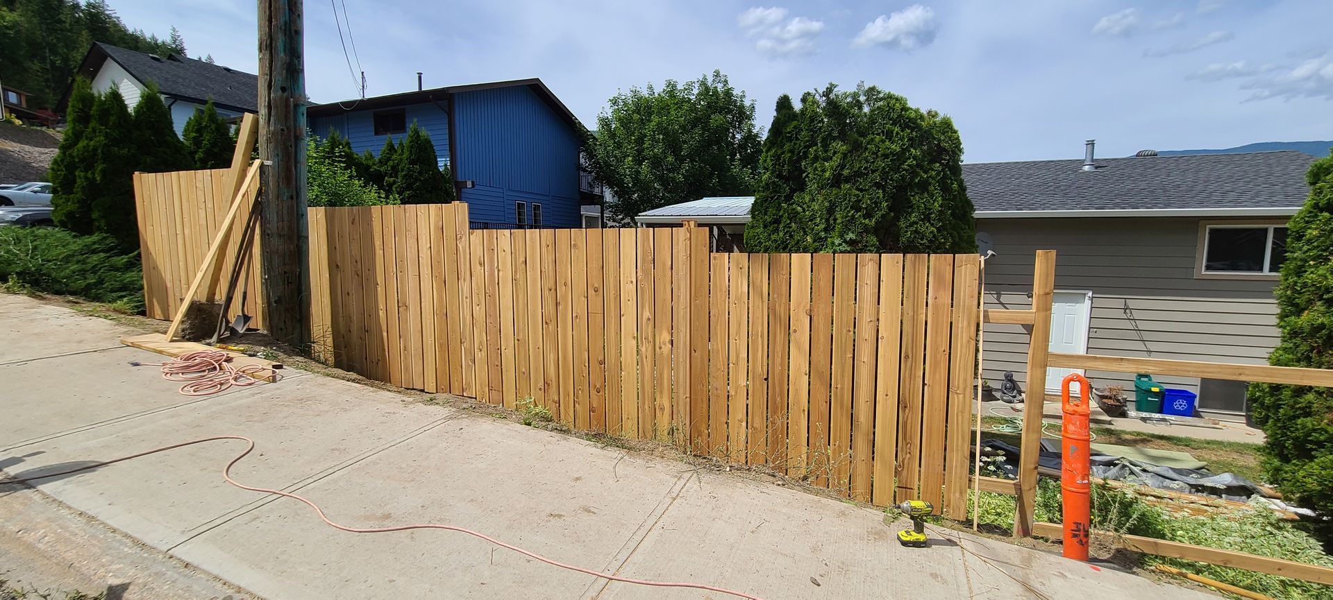 A newly built wooden fence running along a paved road, beside a grassy area. A blue house and green trees are in the background.