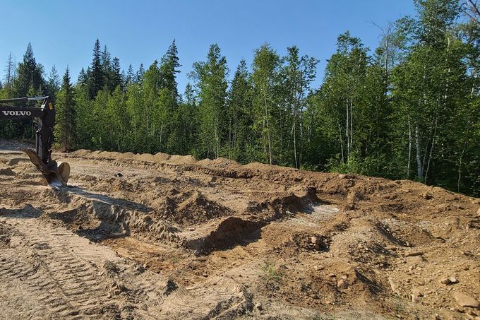 A small excavator digging trenches in dirt with trees in the background on a sunny day.
