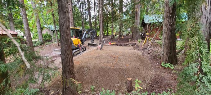 Construction site in a wooded area with machinery, leveled ground, and trees.