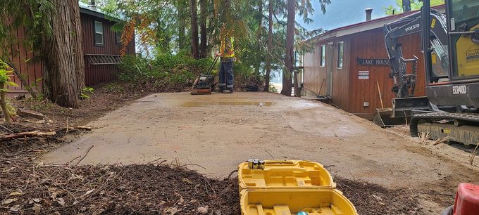 A construction site with a gravel base, trees, and buildings. A yellow tool is in the foreground.