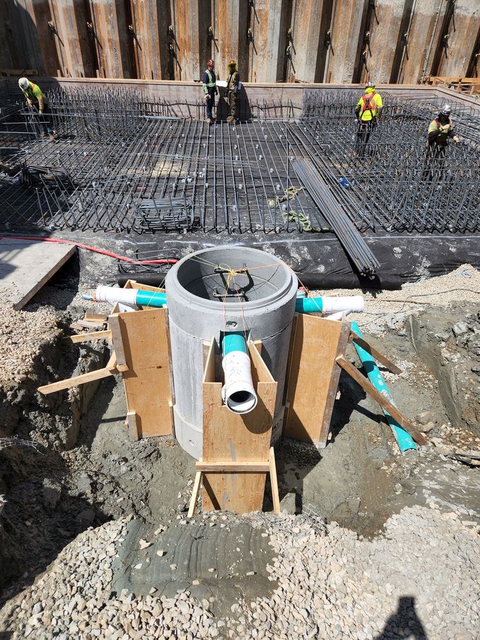 Construction site: concrete drainage structure with connected pipes, workers in the background.
